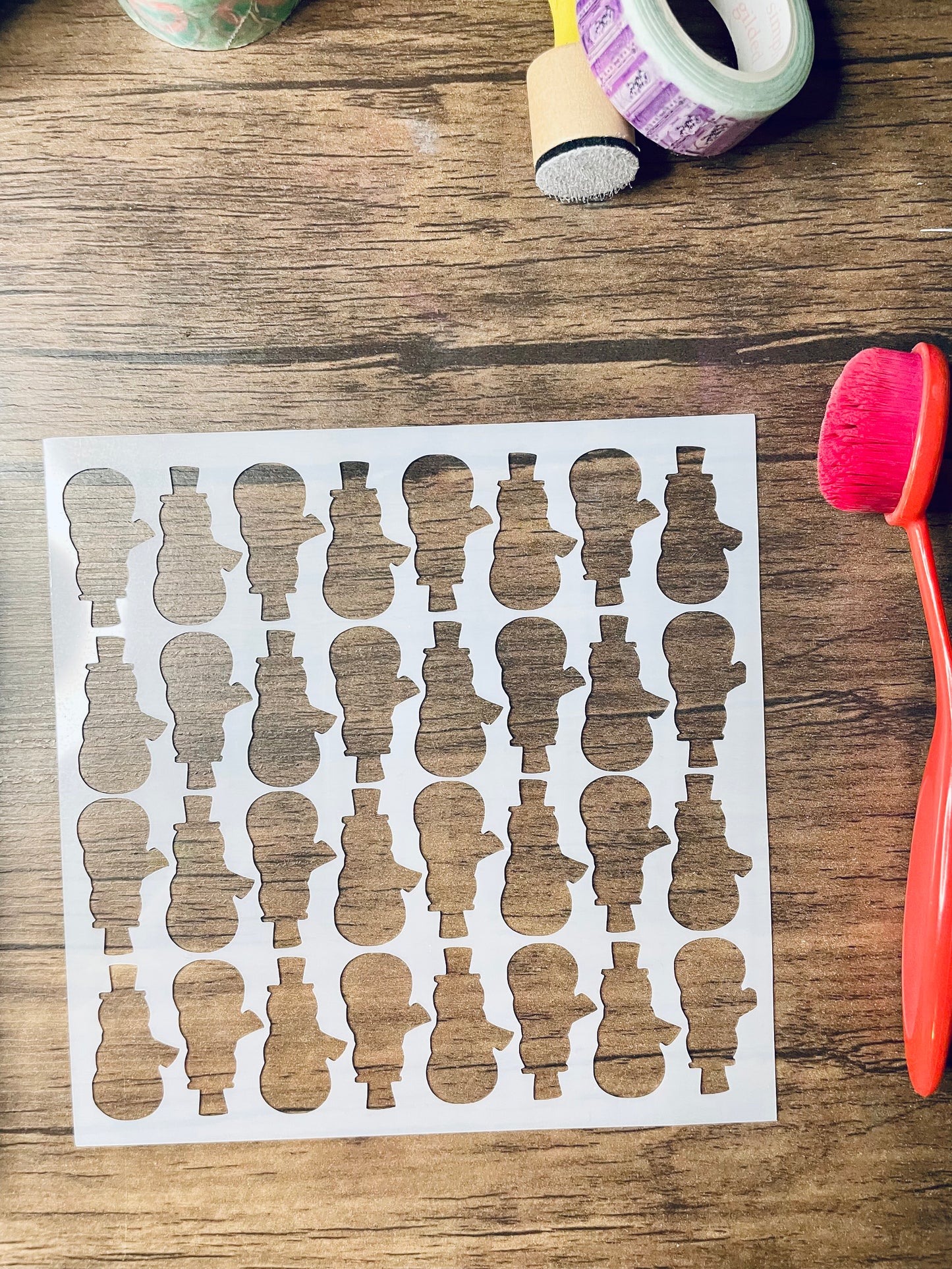 a 5.75"x5.75" stencil sits on a wooden desk. Also on the desk is a red brush and other stationary. The stencil is frosted and includes cutouts of: snowman silhouettes alternating up and down in rows