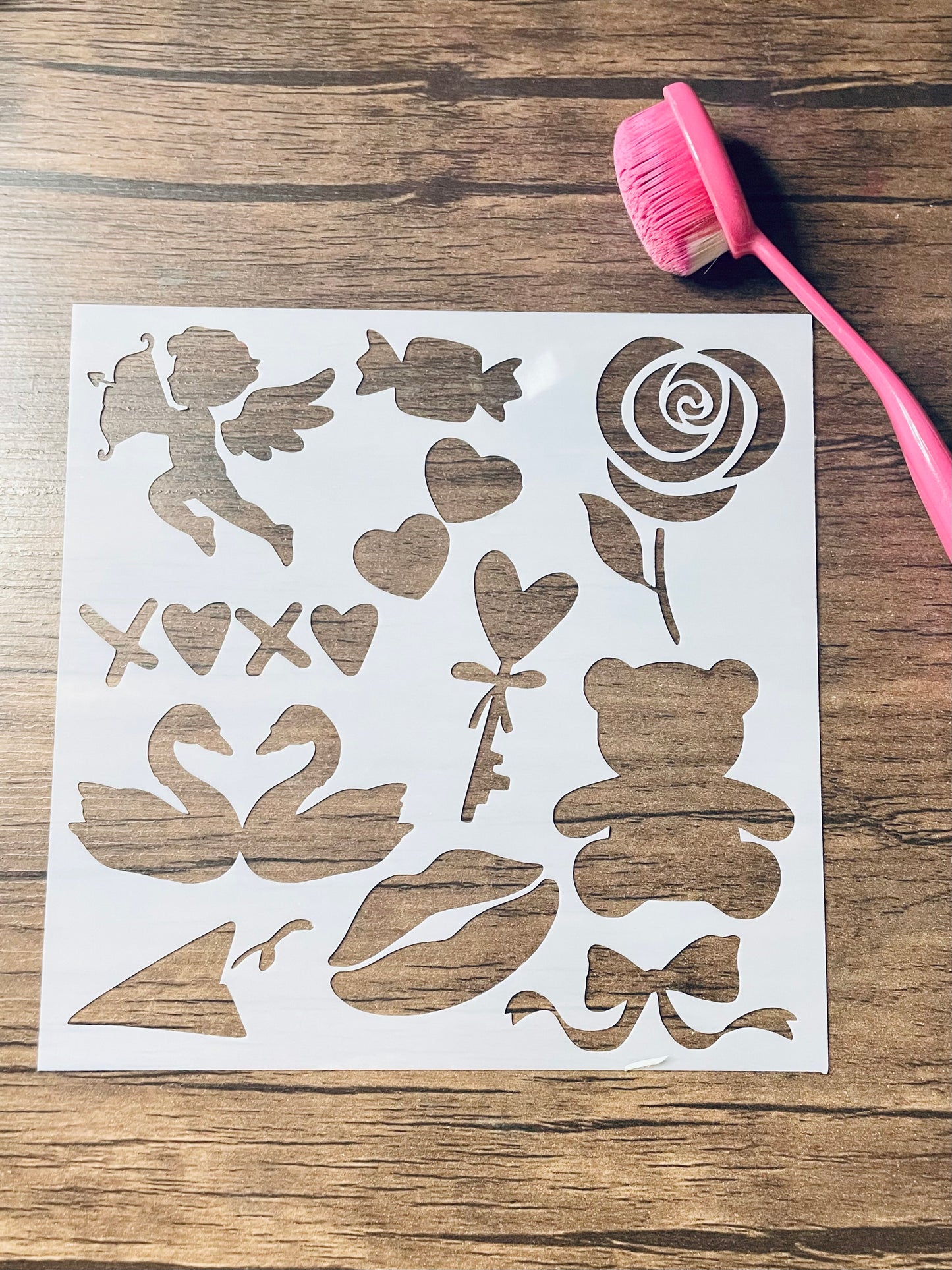 5.75” x 5.75” frosted stencil with Silhouettes of:Cupid, a candy, hearts, a rose, a teddy bear, a heart shaped key, xoxo (o’s are hearts), two swans almost kissing, a paper airplane, a kiss lip, and a bow . Stencil is on a wood surface with a pink brush
