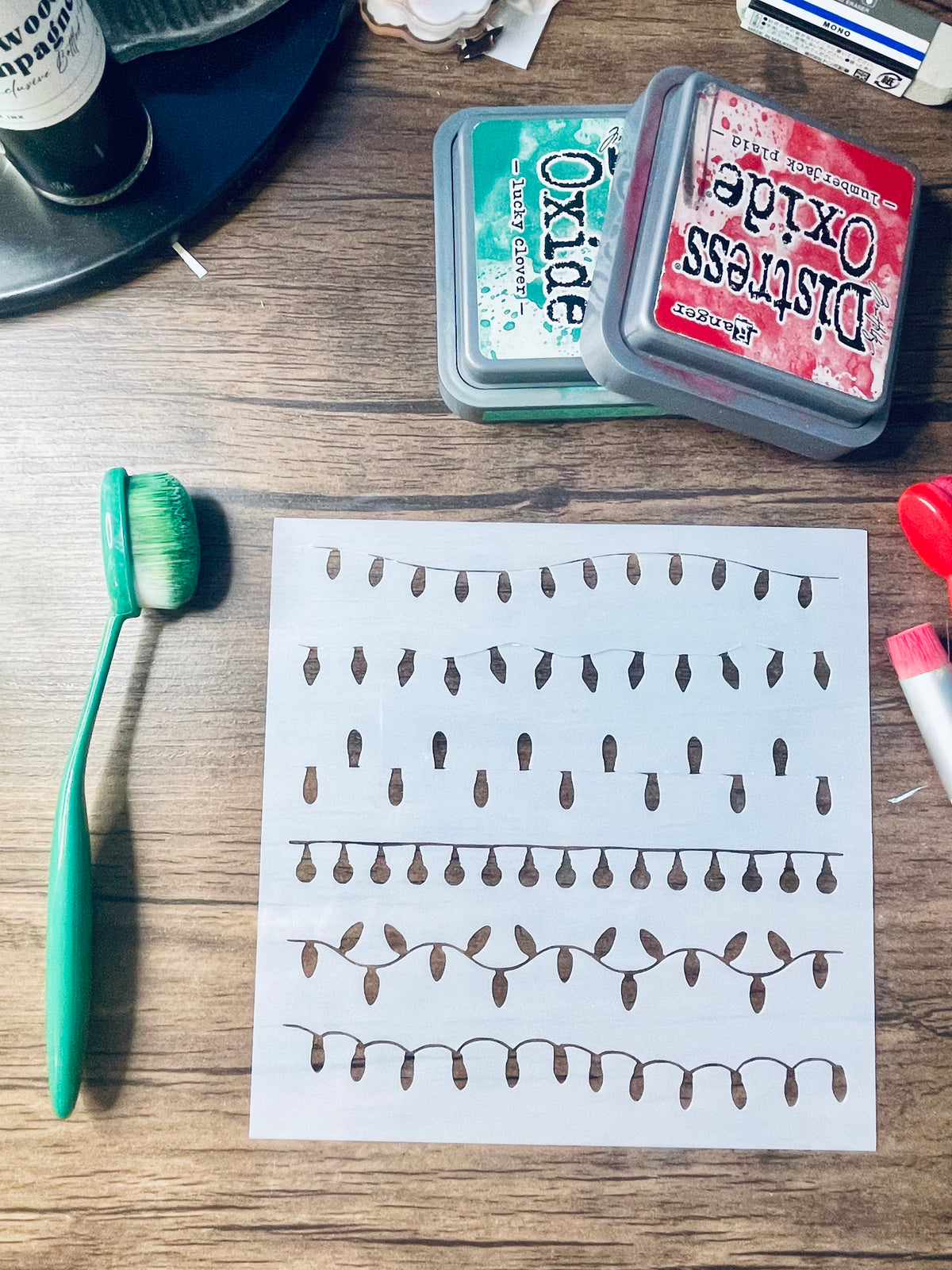 A printed sheet with rows of small brown teardrop-like shapes on white paper, placed on a wooden surface with a green toothbrush, two Distress Oxide ink pads, and other craft supplies nearby.