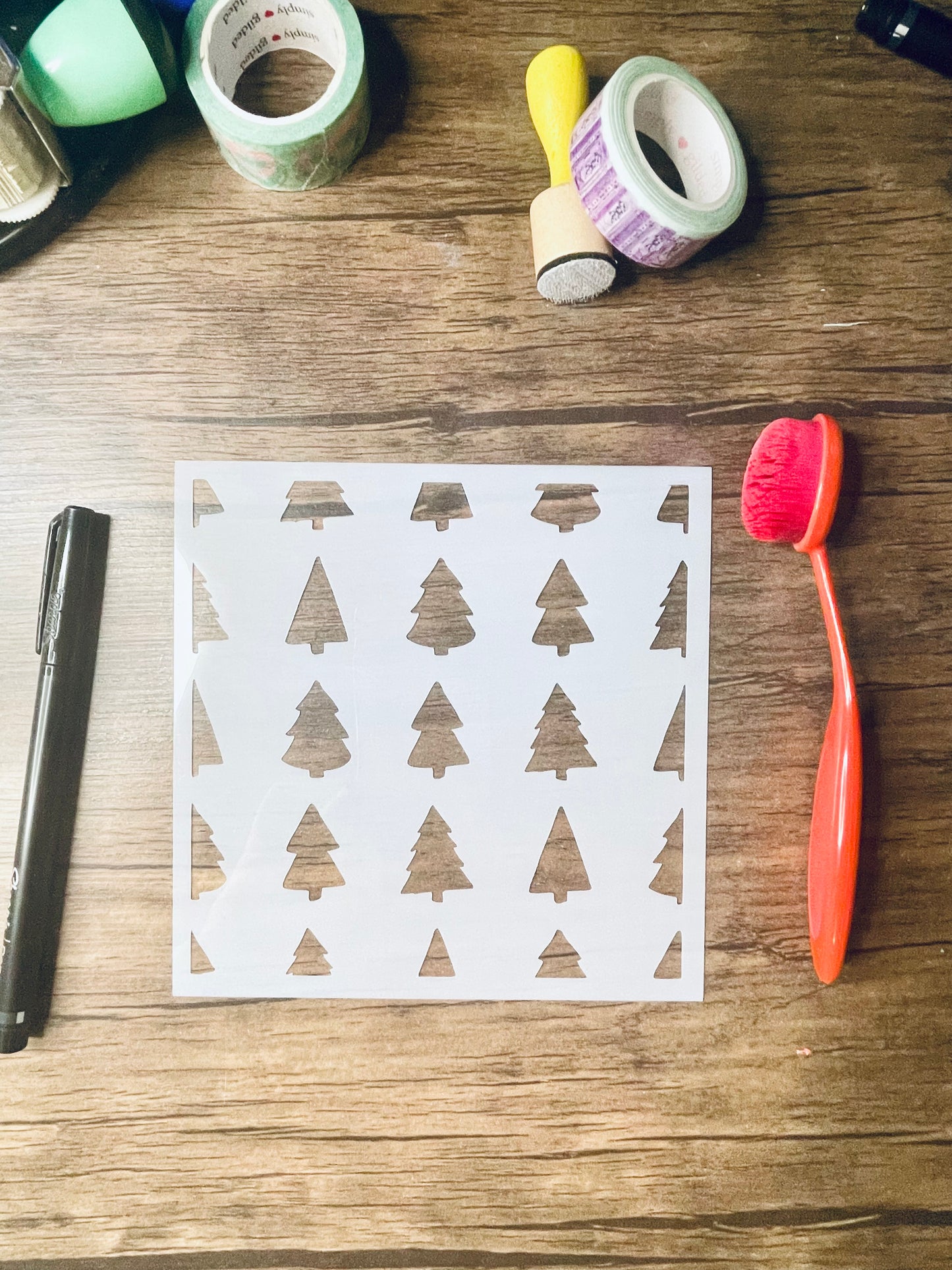 a 5.75"x5.75" stencil sits on a wooden desk. Also on the desk is a red brush and other stationary. The stencil is frosted and includes cutouts of: various shaped christmas trees in a pattern across the stencil