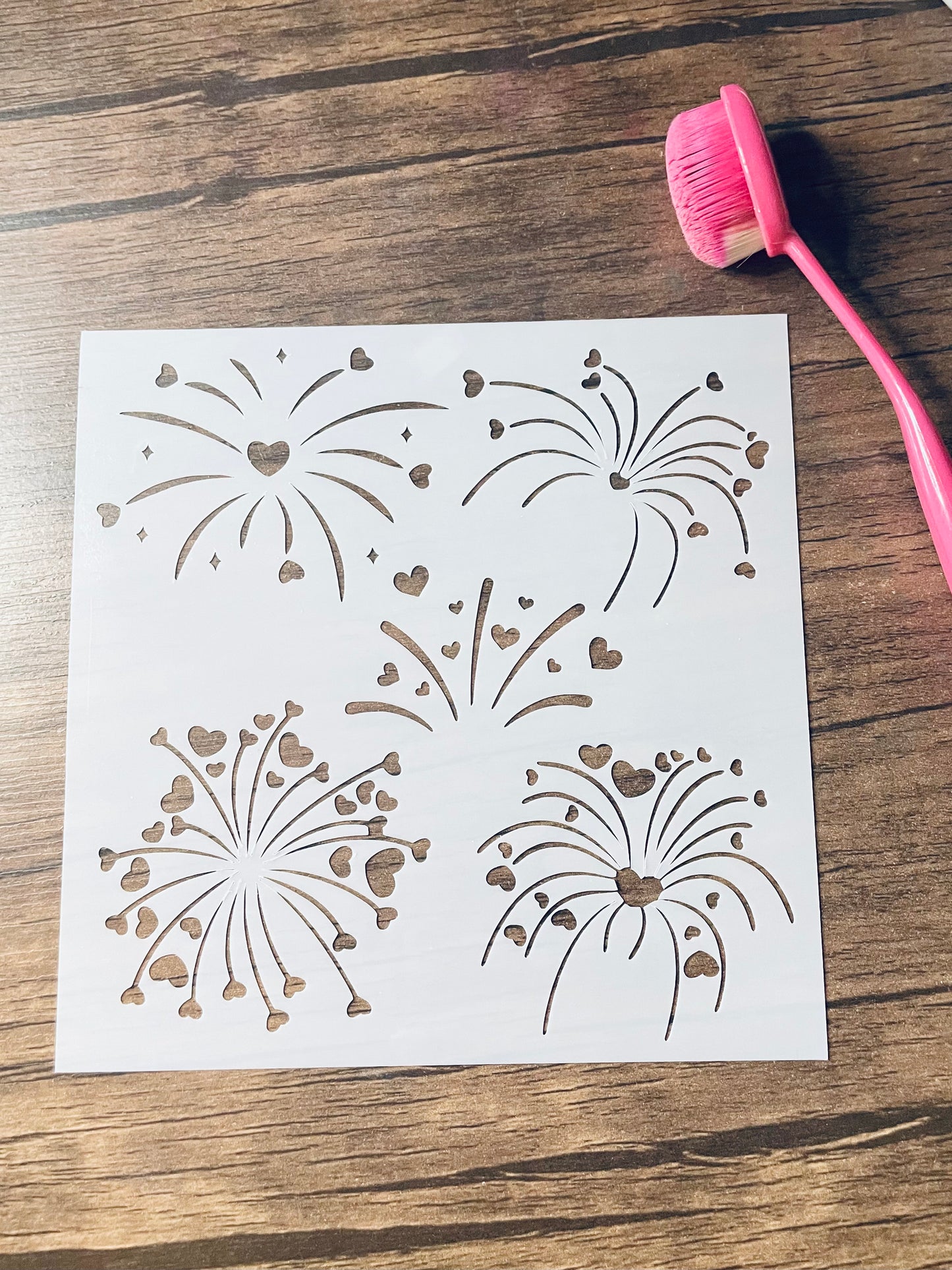 5.75” x 5.75” frosted stencil with Silhouettes of: five different fireworks with heart shaped sparks through them . Stencil is on a wood surface with a pink brush