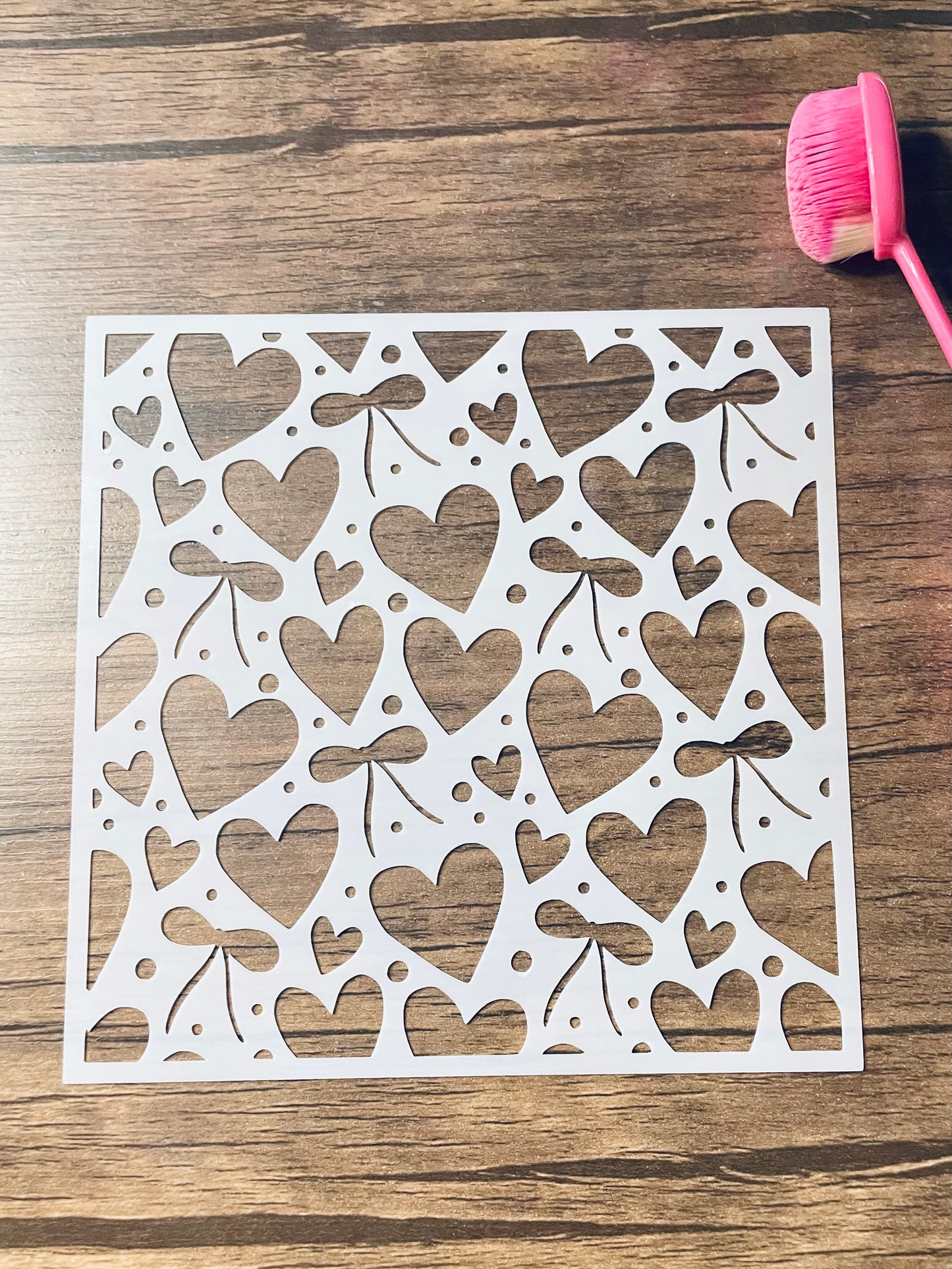 5.75” x 5.75” frosted stencil with Silhouettes of: a variety of hearts bows and dots in a random pattern on the stencil. Stencil is on a wood surface with a pink brush