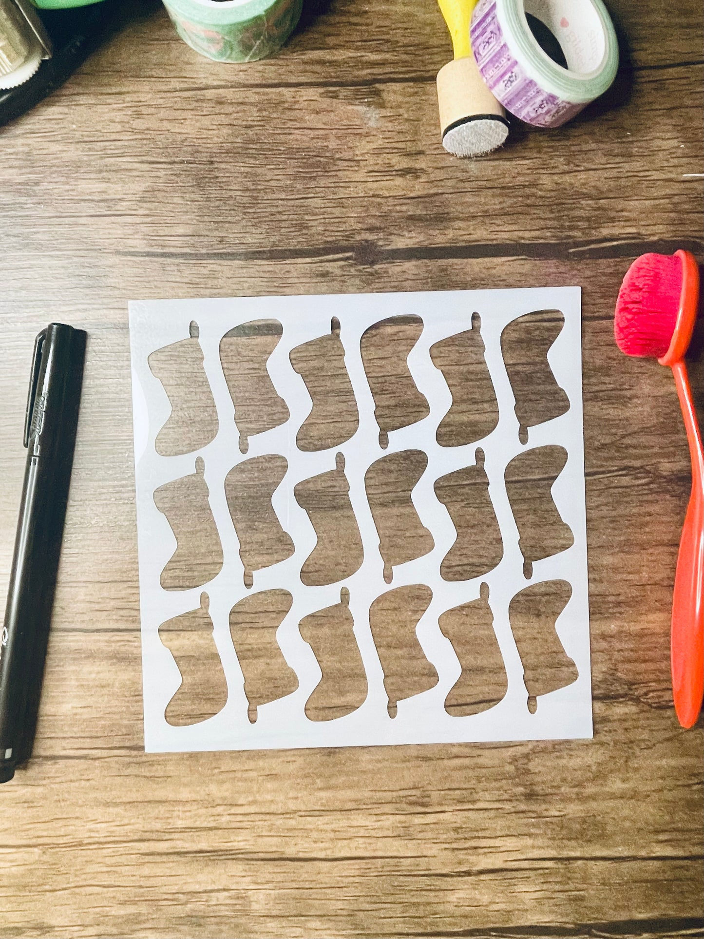 a 5.75"x5.75" stencil sits on a wooden desk. Also on the desk is a red brush and other stationary. The stencil is frosted and includes cutouts of: a stocking in alternating up and down pattern across the stencil. There are three rows of this