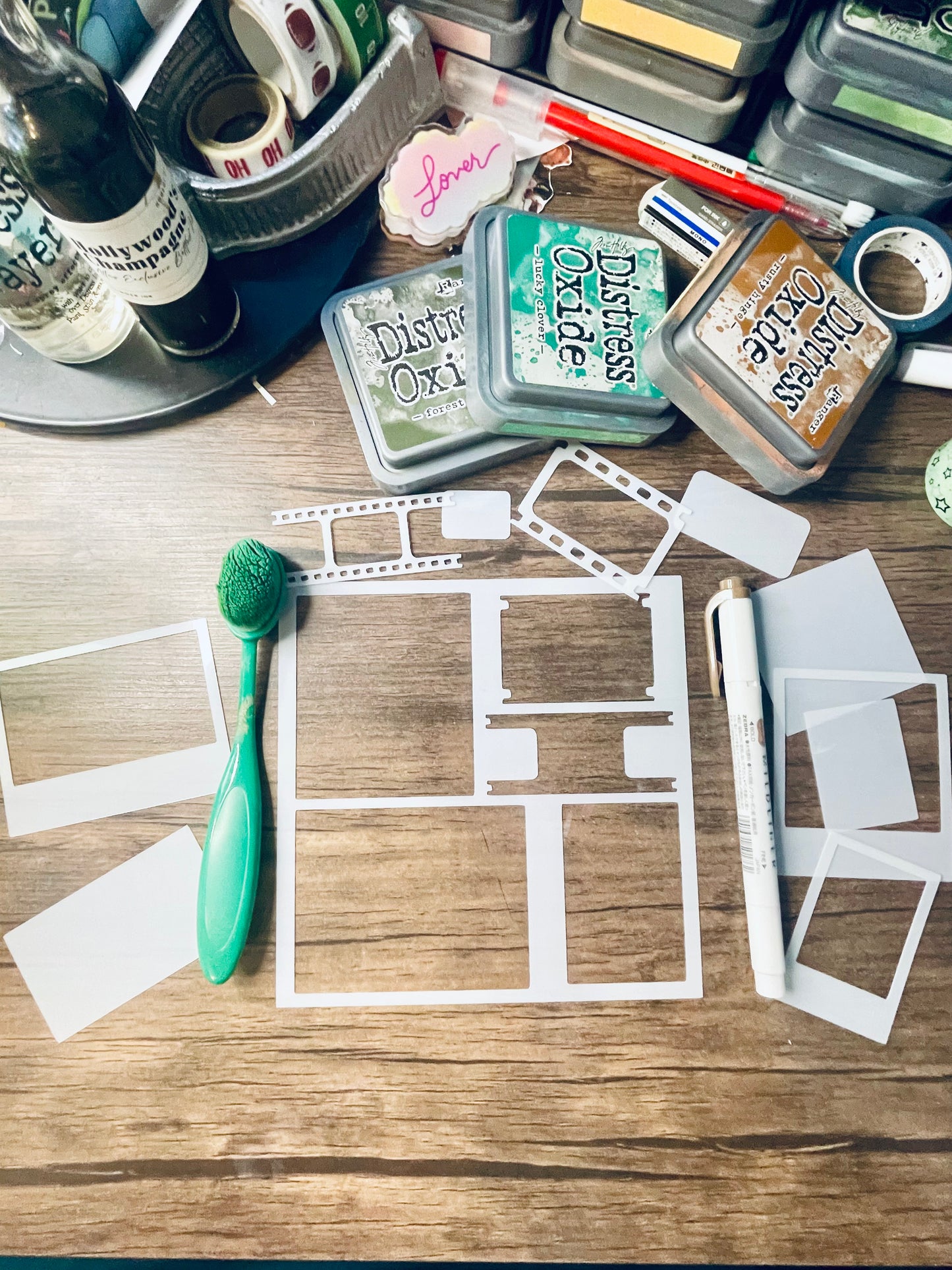 A flat-lay view of a crafting workspace on a wooden table, featuring white die-cut template frames, a green-handled brush, Distress Ink pads, and other craft supplies.