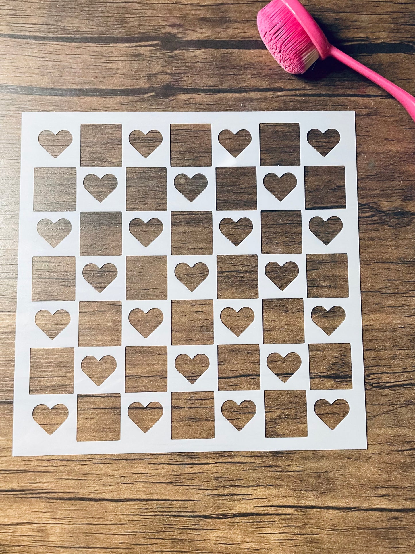 5.75” x 5.75” frosted stencil with cutouts in a checkerboard shape and hearts in alternating squares of the pattern. Stencil is on a wood surface with a pink brush