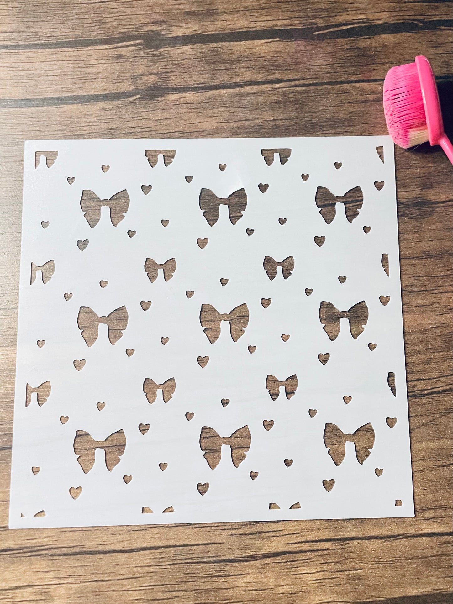 5.75” x 5.75” frosted stencil with Silhouettes of: medium and small bows in a pattern with tiny hearts scattered around. Stencil is on a wood surface with a pink brush