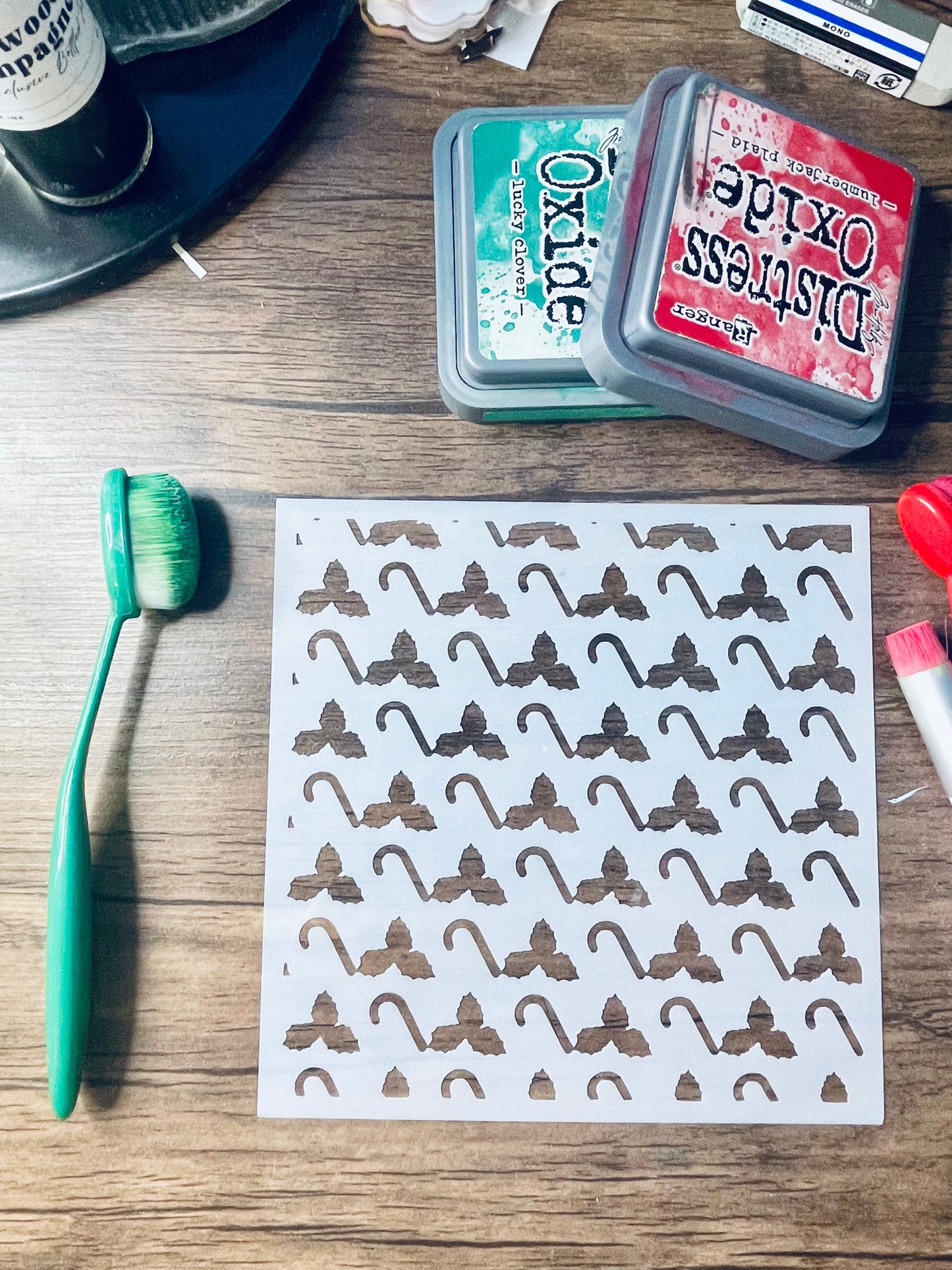 A sheet of white decals or stickers printed with a repeating brown bow-tie pattern, laid on a wooden tabletop alongside a green-handled brush, two Distress Oxide ink pads, and other small craft items.