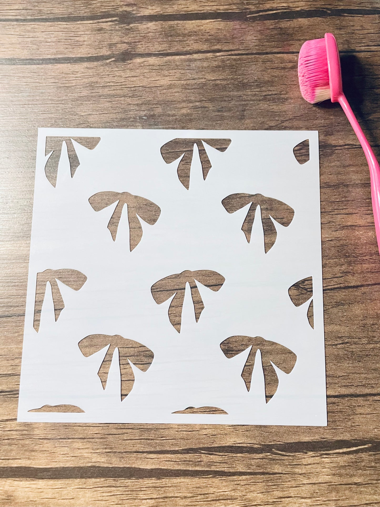 5.75” x 5.75” frosted stencil with Silhouettes of: 5 full bows and multiple parts of bows in a random, seamless pattern. Stencil is on a wood surface with a pink brush