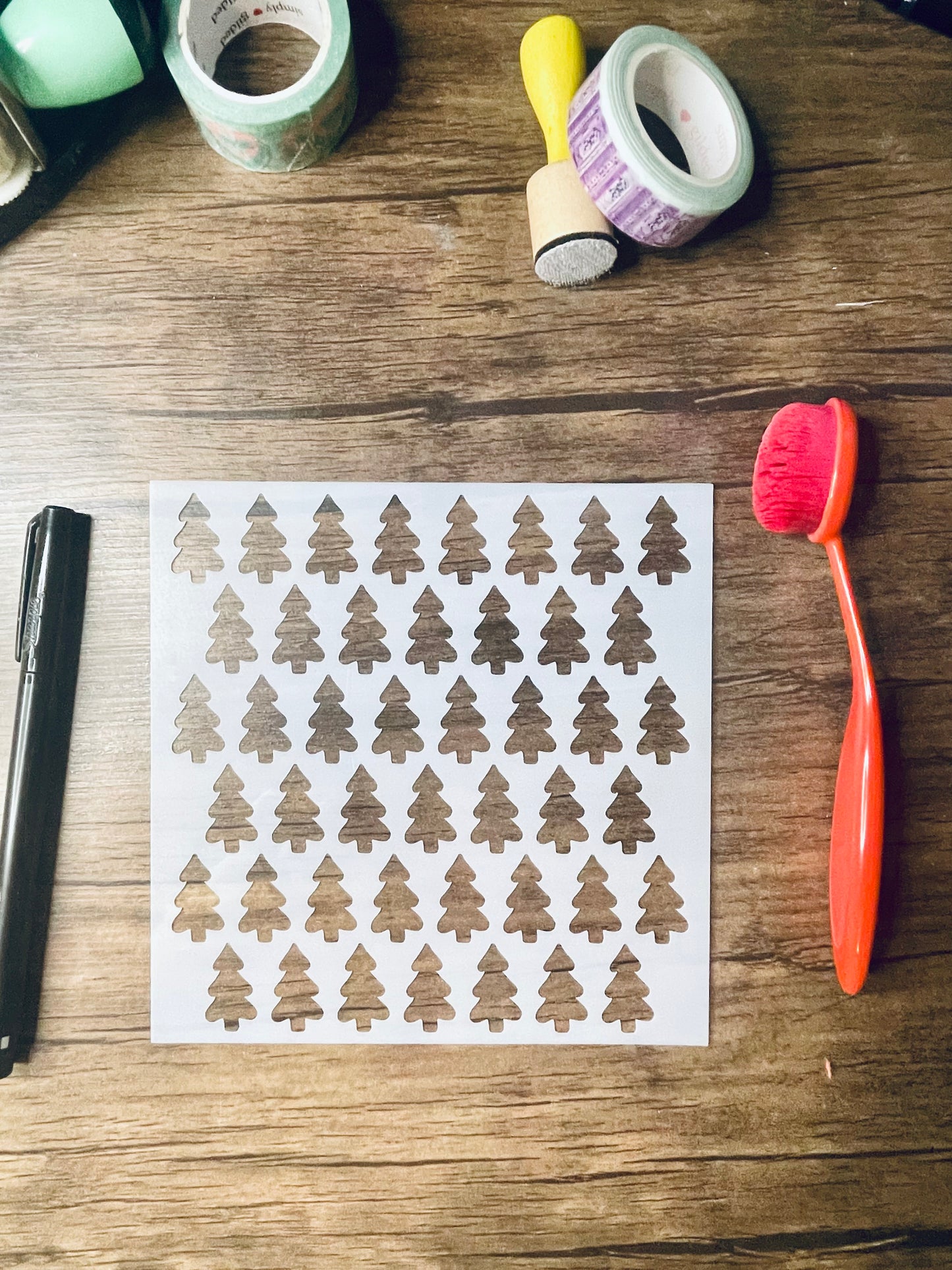 a 5.75"x5.75" stencil sits on a wooden desk. Also on the desk is a red brush and other stationary. The stencil is frosted and includes cutouts of: doodled evergreen style trees in a pattern across the stencil
