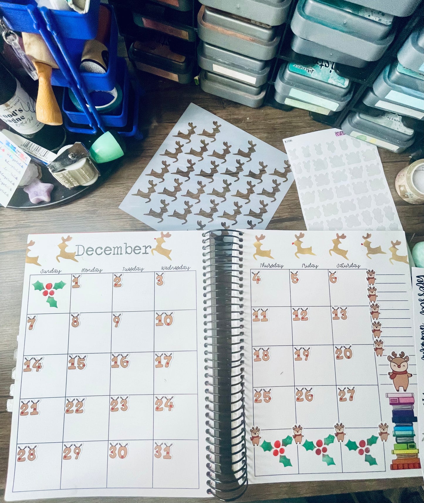 An open planner on a desk. There is also reindeer stencil, vaerious stationary items on the desk. Planner is a december monthly layout decorated with reindeer numbers and reindeer stenciling