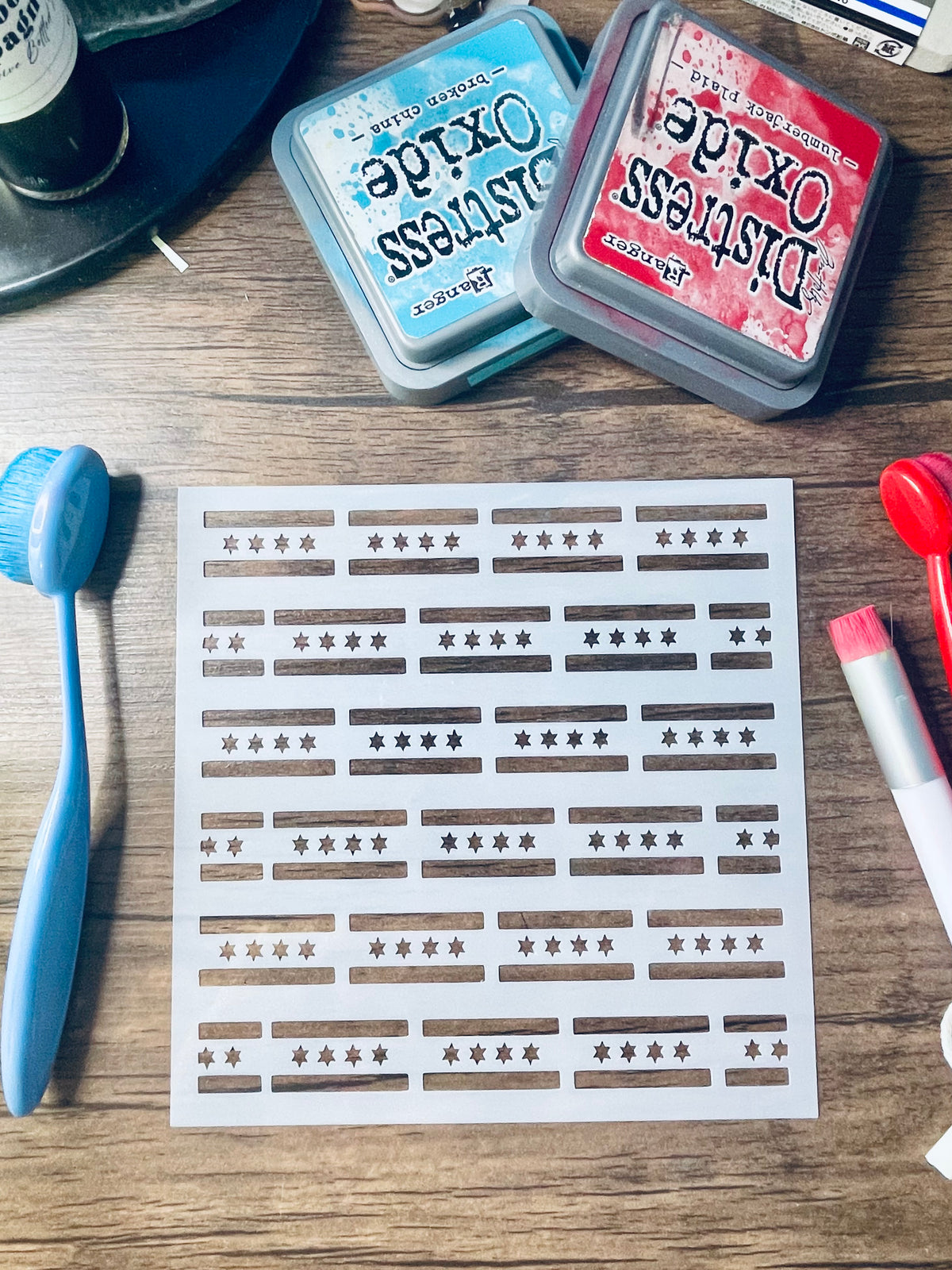 A sheet of nail wraps or stickers with horizontal brown bars and star motifs, displayed on a wooden table alongside Distress Oxide ink pads, a blue brush, and other craft tools.