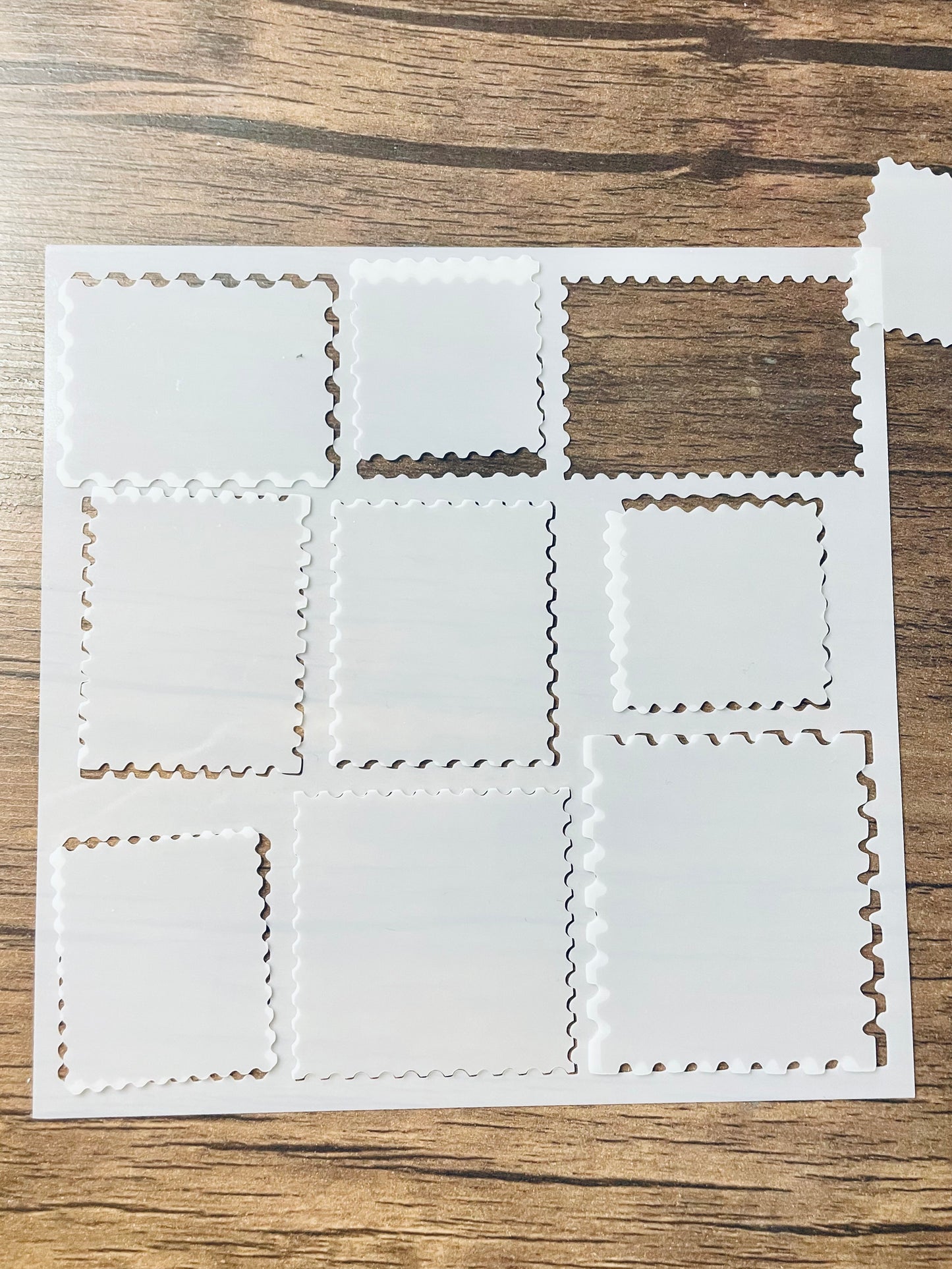 5.75” x 5.75” frosted stencil with Silhouettes of: 9 different stamp shaped cutouts. The cutout pieces are loosely in their cut out areas, other than one piece which is to the side. Stencil is on a wood surface with a pink brush