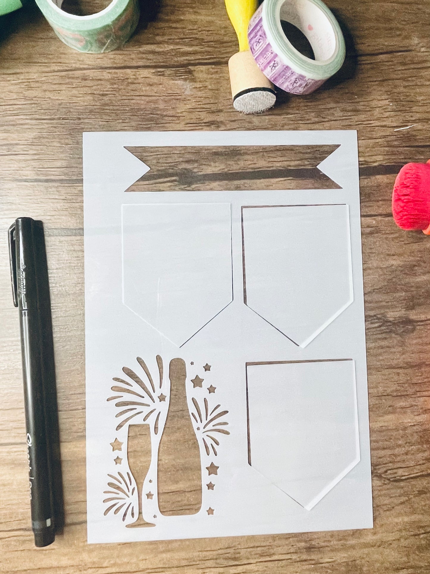 a rectangular stencil sits on a wooden desk. Also on the desk is a red brush and other stationary. The stencil is frosted and includes cutouts of: a banner on top, a row of 2 pentagon shaped banners, followed by one more on the bottom. next to bottom banner is a champagne bottle, champagne flute, and fireworks and stars behind them. The cutouts of the 3 pentagon shaped stencils are loosely in their cut out area