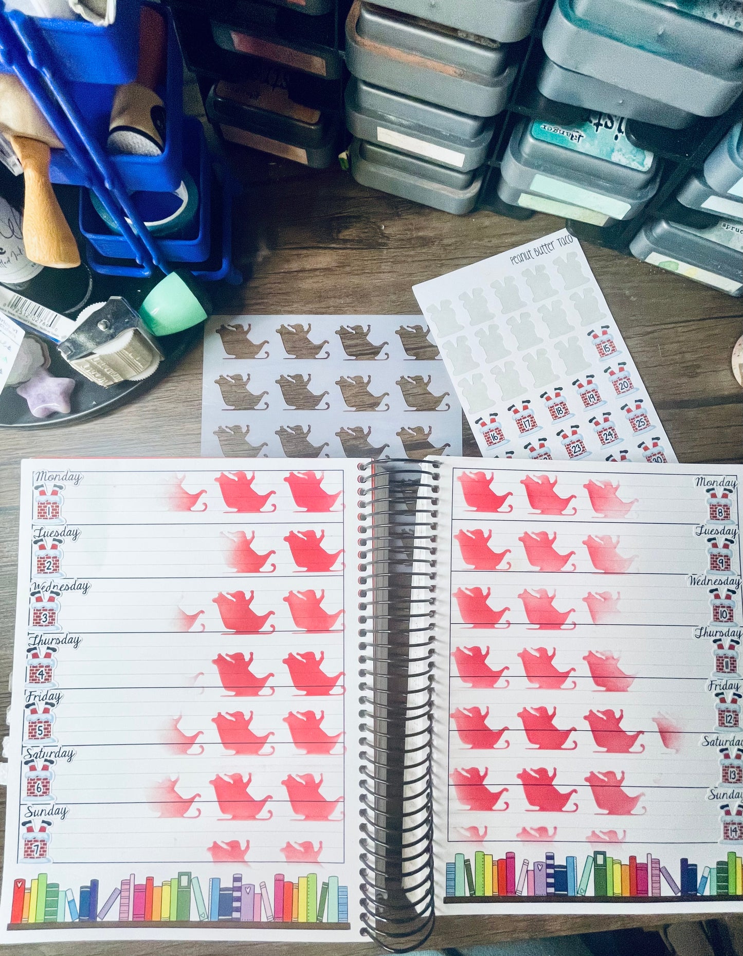 Open planner on a desk with christmas themed numbers and an inked sleigh pattern on it. Wooden desk also has sleigh stencil and a sticker sheet of christmas themed numbers