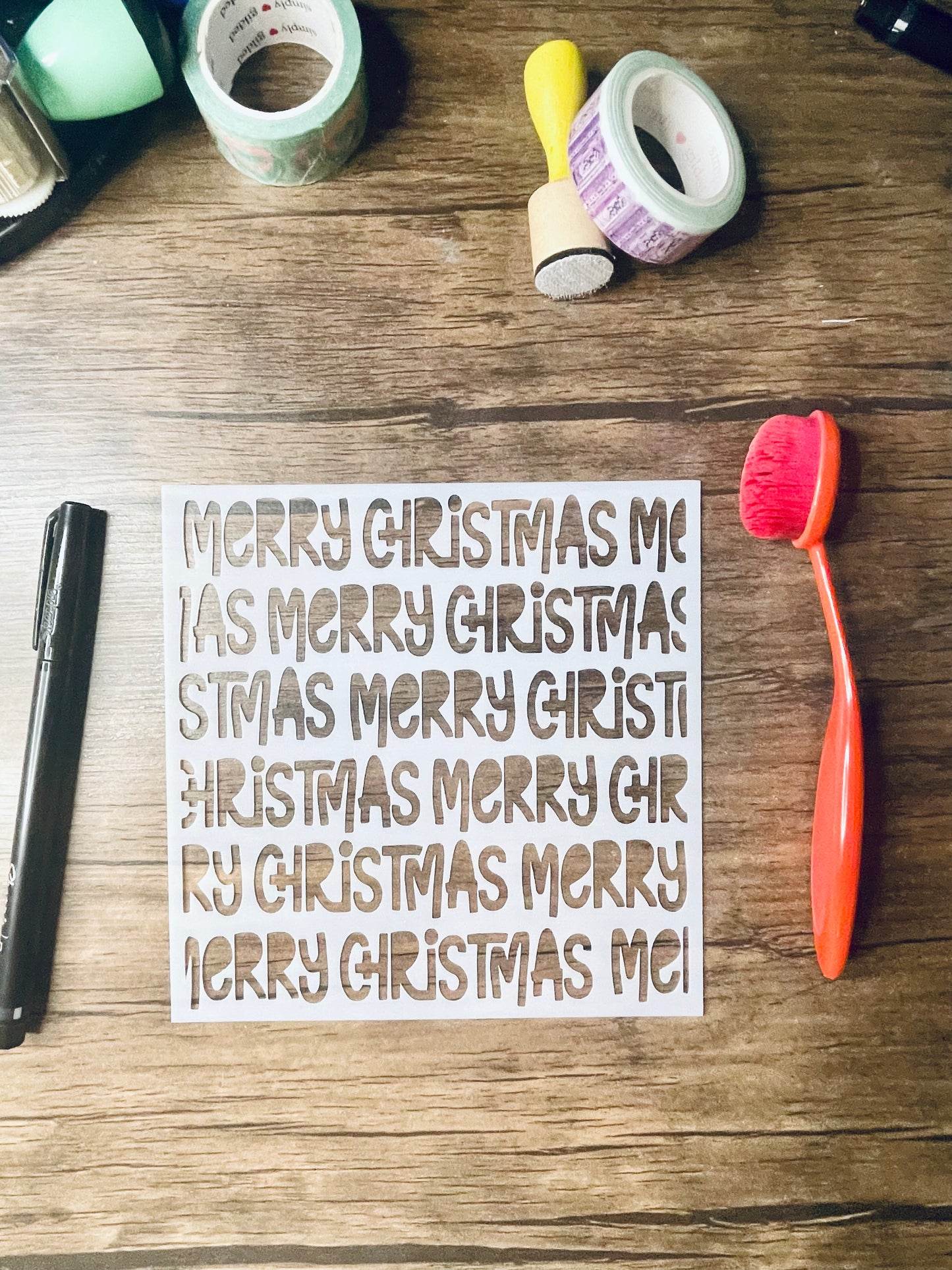 a 5.75"x5.75" stencil sits on a wooden desk. Also on the desk is a red brush and other stationary. The stencil is frosted and includes cutouts of: "merry christmas" in horizontal lines in a pattern