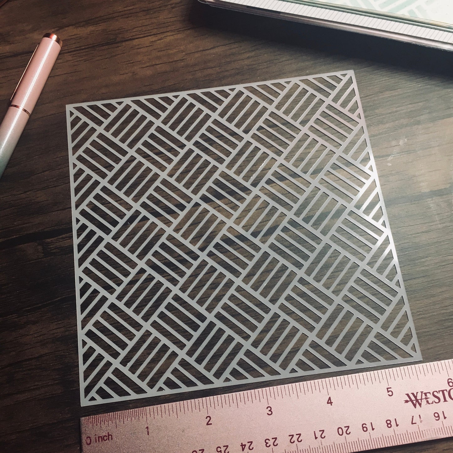 A stencil with a geometric pattern is laying on a wooden surface next to a ruler, pen and notebook