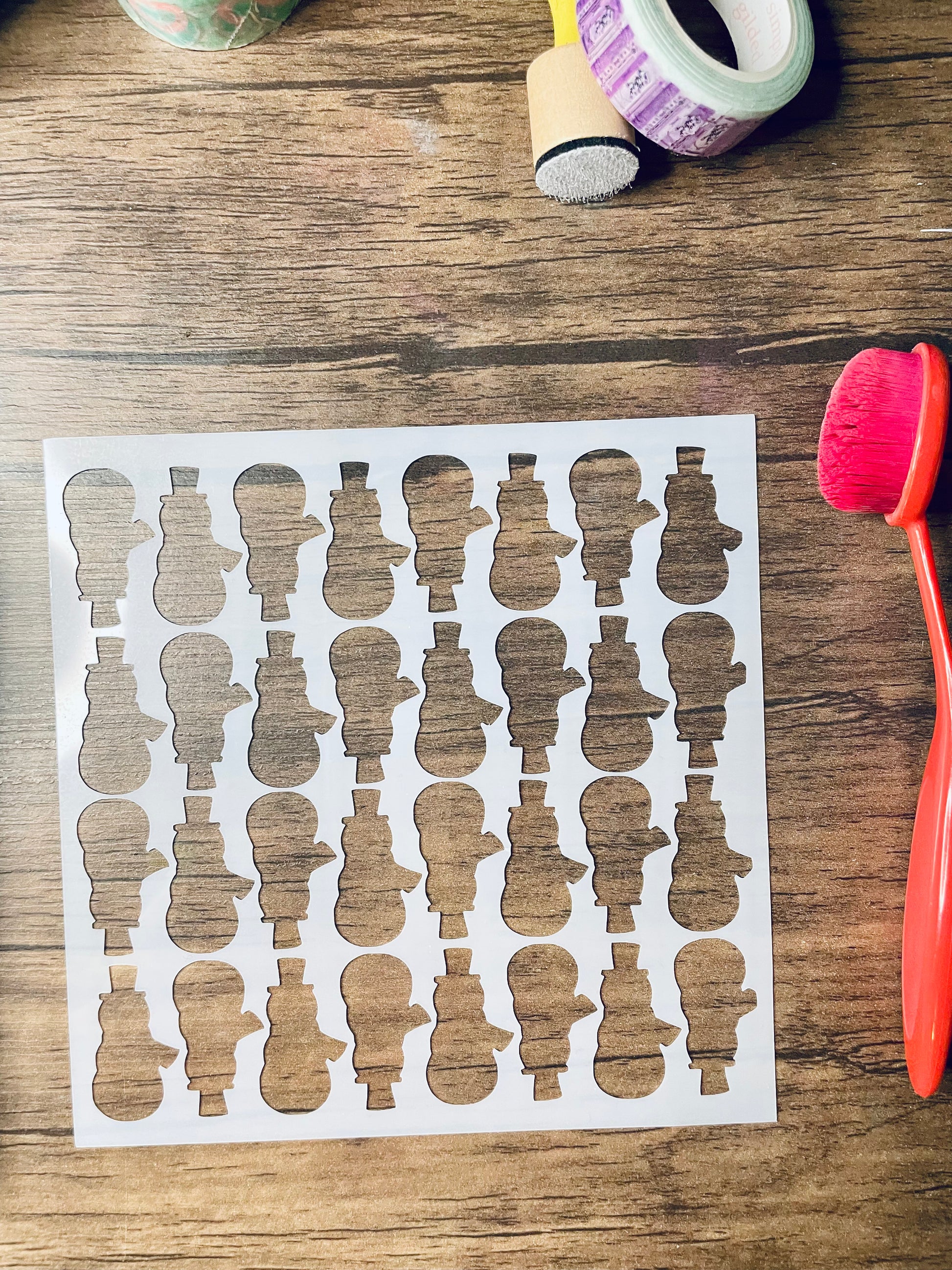 a 5.75"x5.75" stencil sits on a wooden desk.  Also on the desk is a red brush and other stationary.  The stencil is frosted and includes cutouts of: snowman silhouettes alternating up and down in rows 