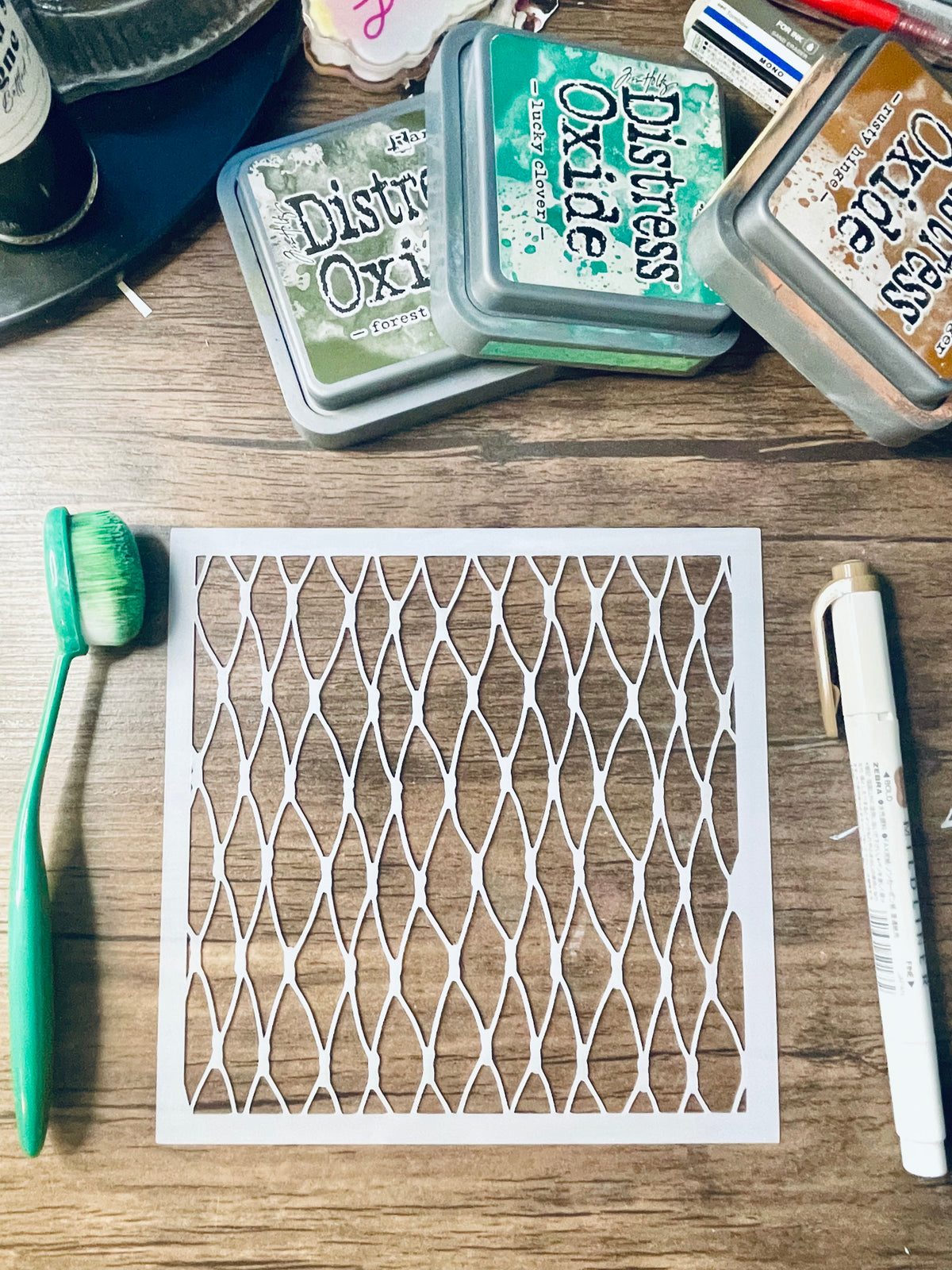 A square white stencil with a repeating diamond lattice pattern laid flat on a wooden surface, surrounded by craft supplies including Distress Oxi ink pads, a green toothbrush, and a white marker.