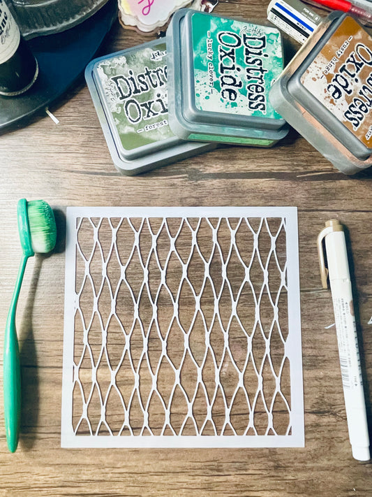 A square white stencil with a repeating diamond lattice pattern laid flat on a wooden surface, surrounded by craft supplies including Distress Oxi ink pads, a green toothbrush, and a white marker.