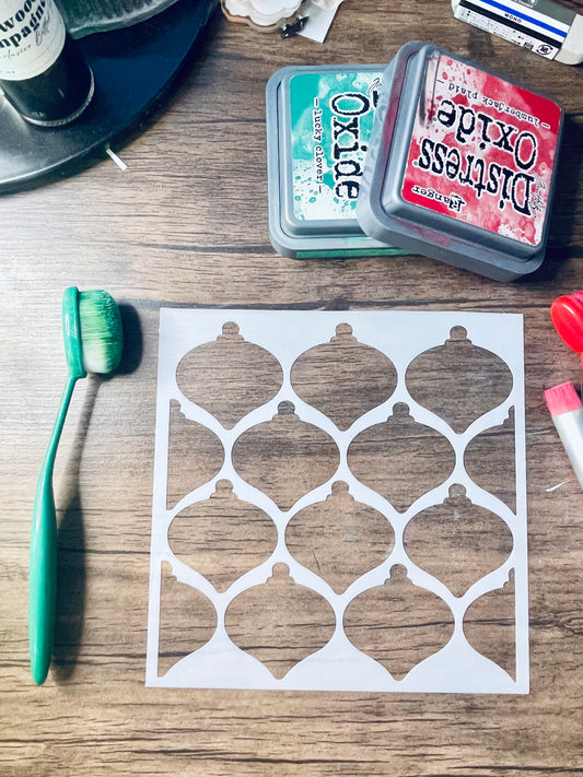 A white stenciled ornament pattern sheet displayed on a wooden surface, surrounded by craft supplies including a green-handled brush, an Oxide ink pad, and other crafting tools.