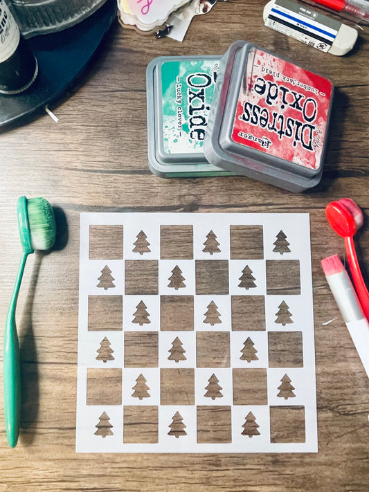 A square printed sheet with a white and brown checkerboard pattern and tree-shaped cutouts, arranged on a wooden tabletop with craft tools and ink pads nearby.