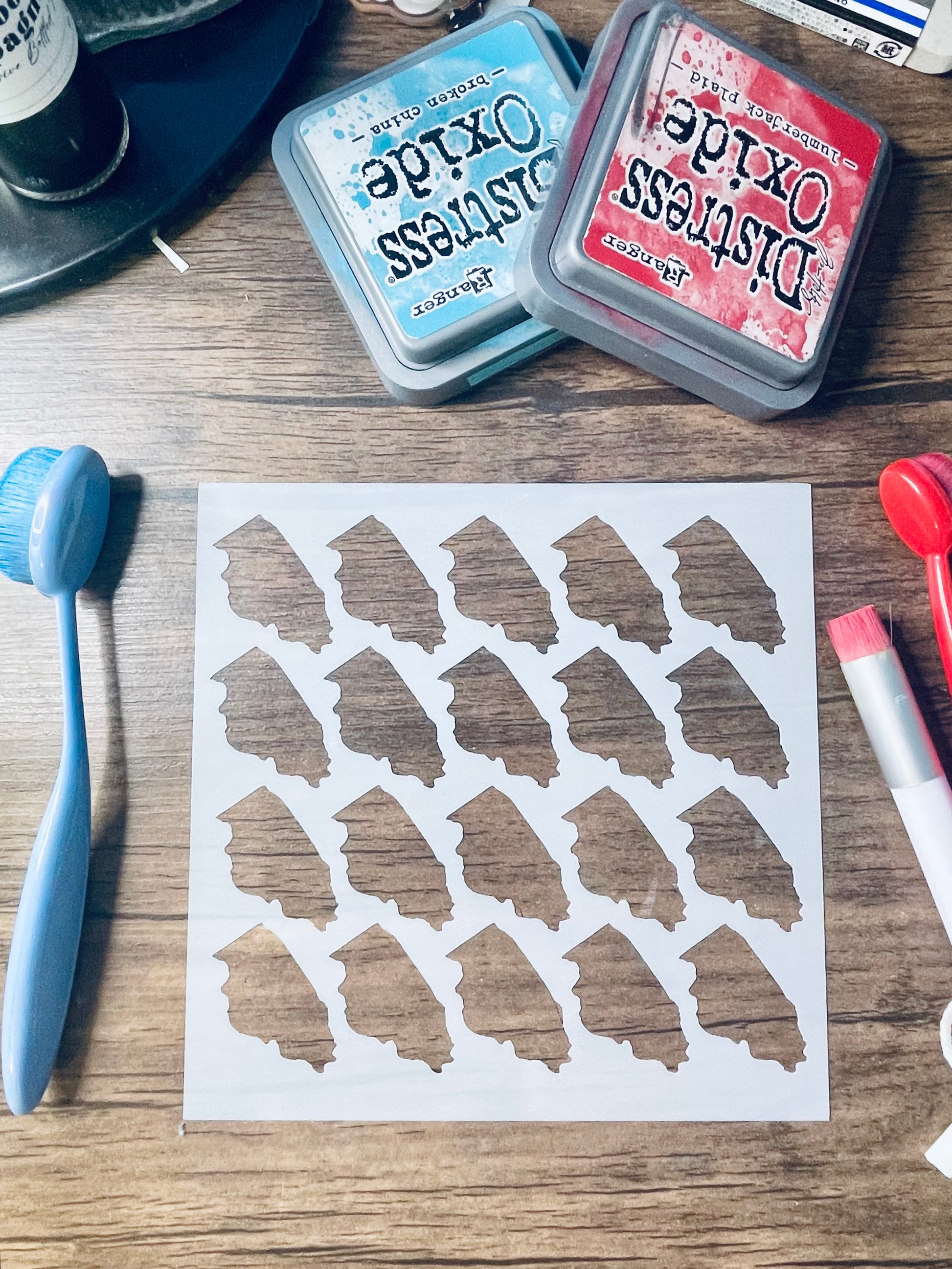 A sheet of white paper with multiple small irregular brown shapes stamped or printed on it, arranged on a wooden tabletop. Two Distress Oxide ink pads, a blue sponge, and other craft tools are visible around it.