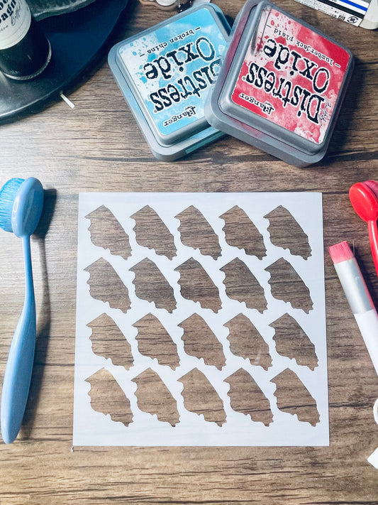 A sheet of white paper with multiple small irregular brown shapes stamped or printed on it, arranged on a wooden tabletop. Two Distress Oxide ink pads, a blue sponge, and other craft tools are visible around it.