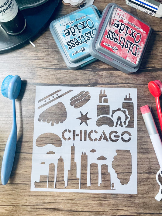 A white stenciled paper sheet featuring Chicago skyline silhouettes and the word CHICAGO in the center, laid on a wood surface with craft tools and two Distress Oxide ink pads around it.