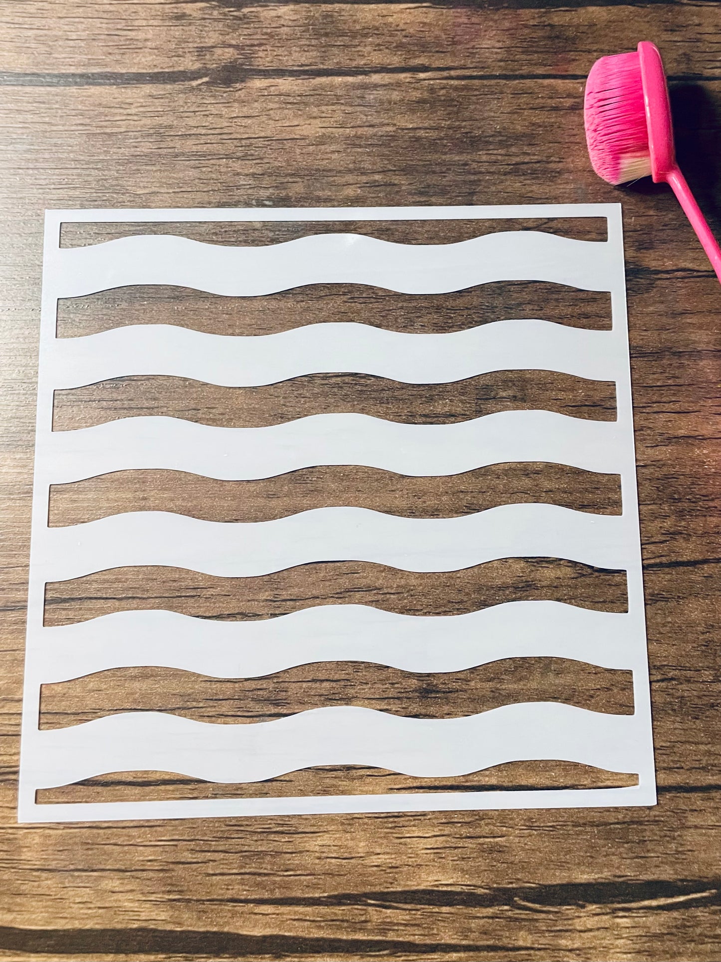 5.75” x 5.75” frosted stencil with Silhouettes of: six horizontal wavy stripes.    Stencil is on a wood surface with a pink brush 