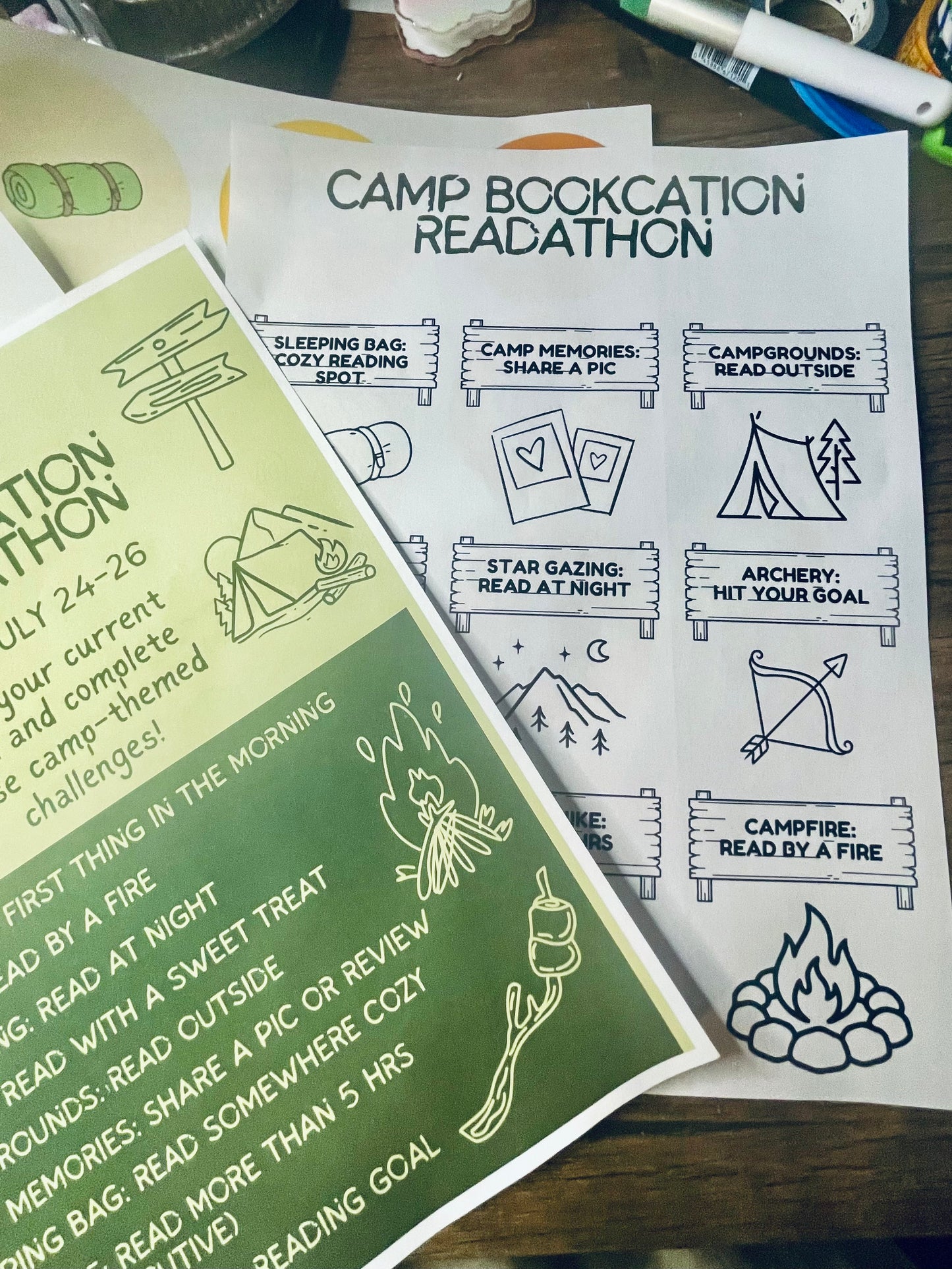 Two printed reading challenge worksheets laid on a wooden desk, one green and one white, with camp-themed illustrations and text such as 'Camp Bookathon Readathon.'.