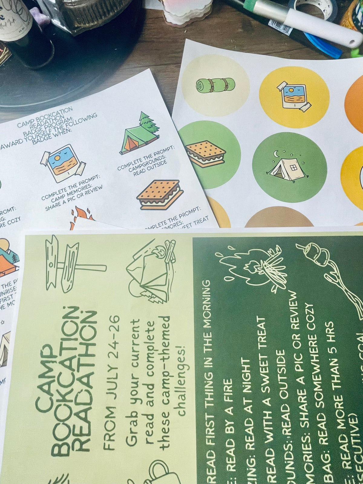A close-up of printed reading challenge and sticker sheets laid on a desk, with visible books and stationery in the background. The sheets contain illustrated books, a tent, a campfire, and text prompts such as 'Camp Bookcation Readathon' and 'From July 24-26'.