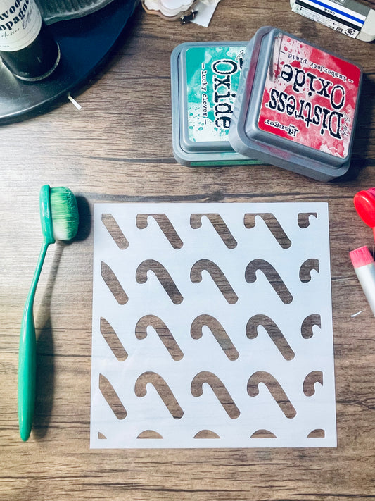 A white square stencil sheet with repeated curved cutout shapes, laid on a wooden surface. A green-handled brush is placed to the left, and two Distress Oxide ink pads are visible at the top right.