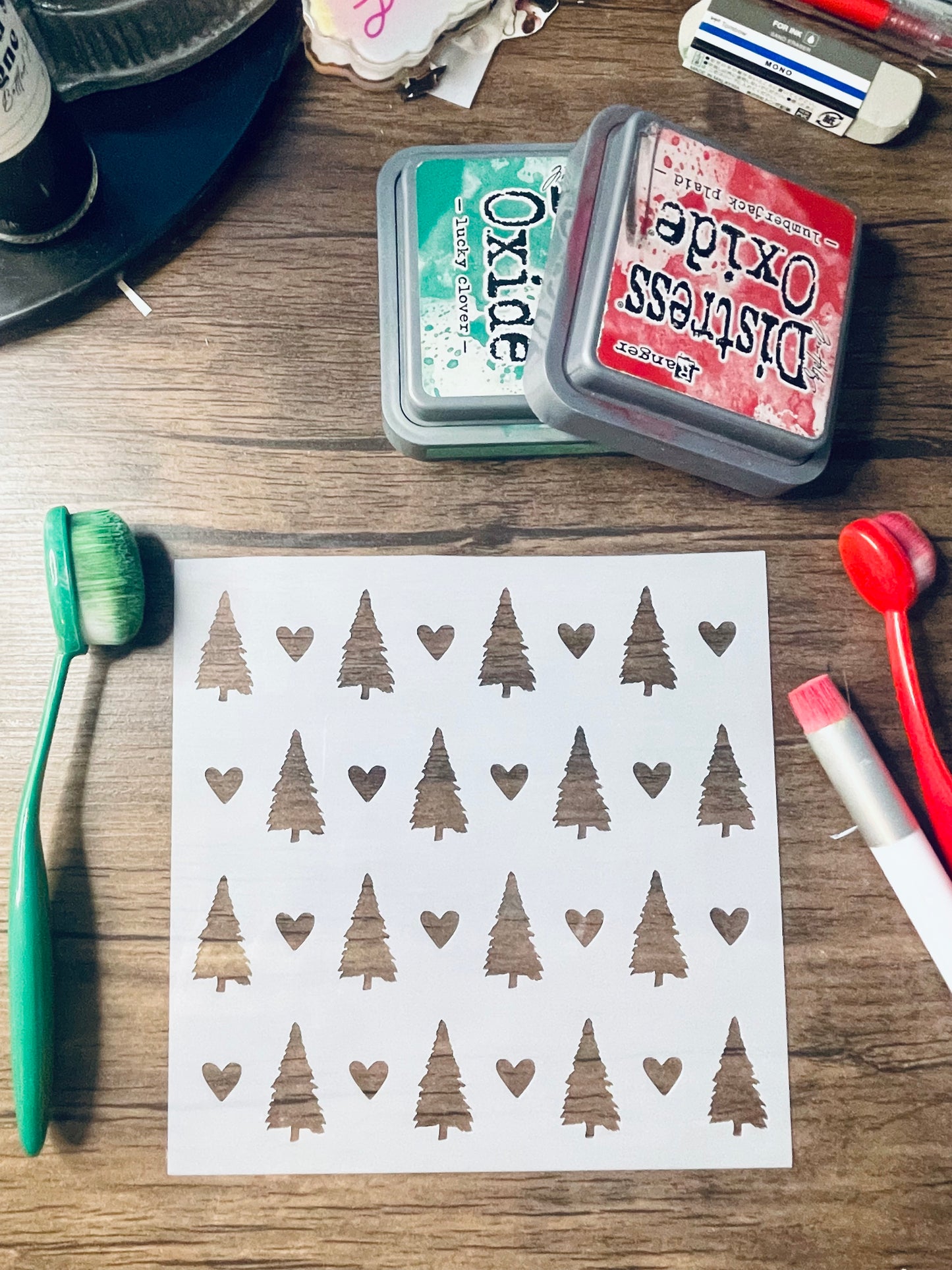 A sheet of white transfer stickers featuring a repeating pattern of brown stamped Christmas trees and hearts, displayed on a wooden desk alongside craft tools including an Oxide ink pad, a green toothbrush, and a red-handled brush.
