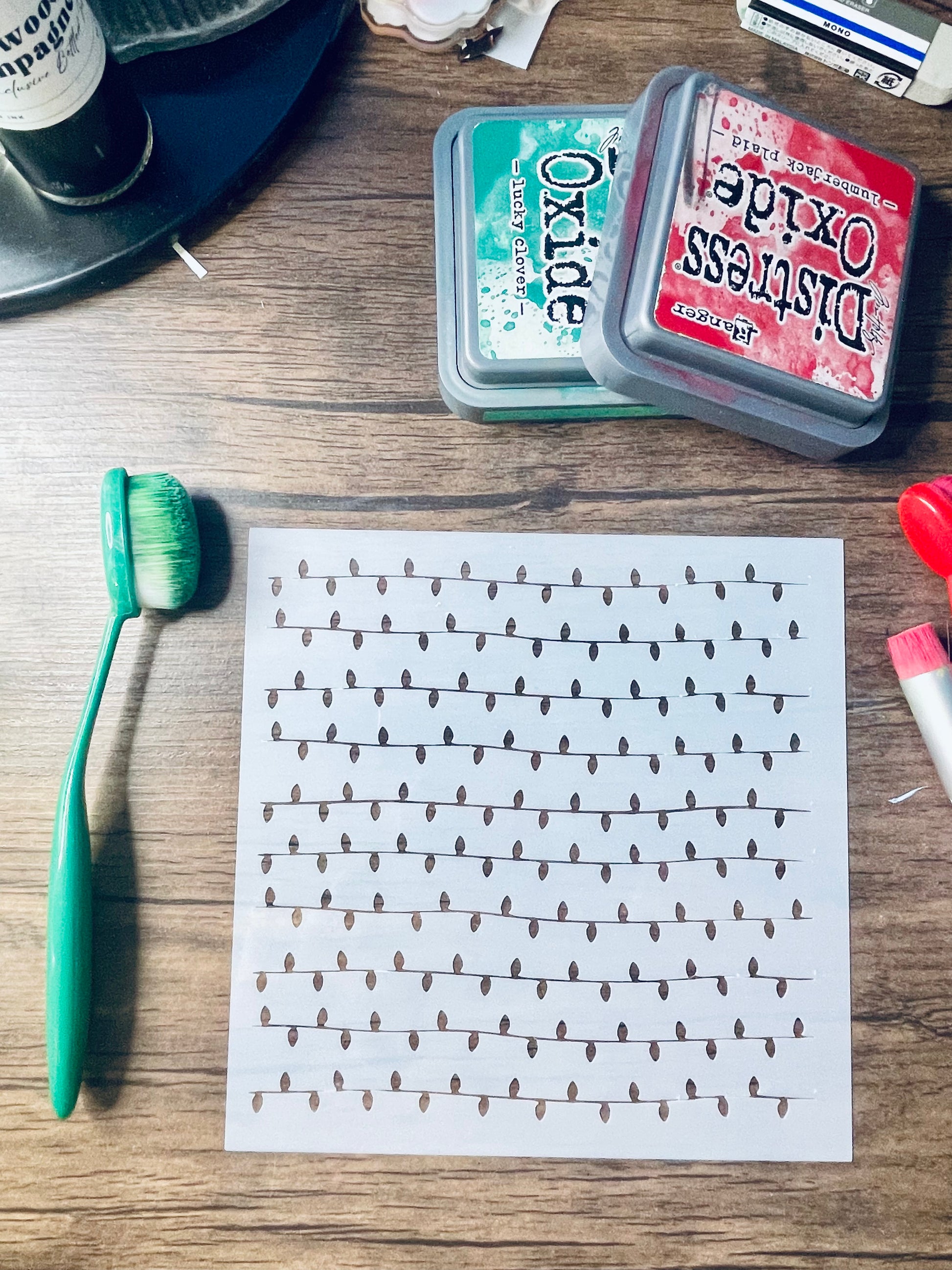 A printed sheet with rows of small black teardrop-like marks on a white background, placed on a wooden tabletop alongside a green toothbrush, two Distress Oxide ink pads, and other craft supplies.