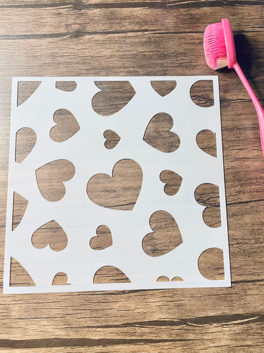 5.75” x 5.75” frosted stencil with Silhouettes of: a variety of sized hearts rotated in a random pattern.   Stencil is on a wood surface with a pink brush 