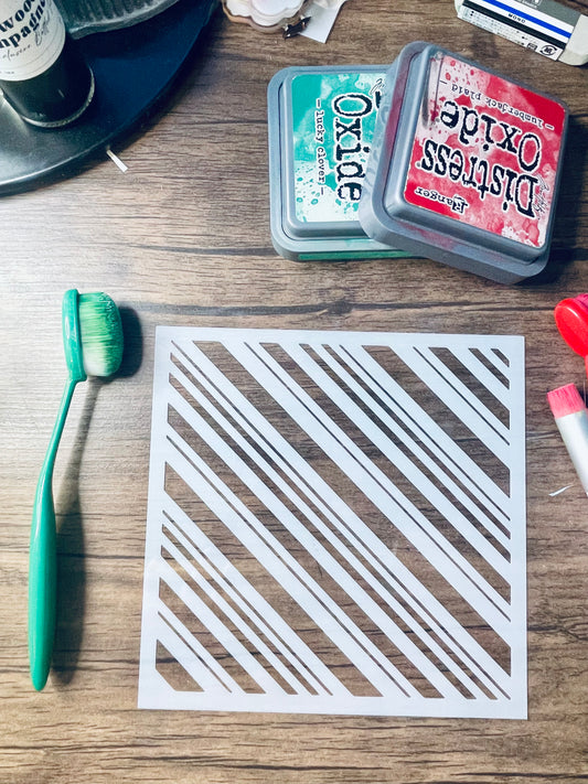 A square white stencil sheet with diagonal striped cutouts lies on a wooden tabletop, surrounded by craft supplies including a green toothbrush, an Oxide ink pad, a Distress Ink pad, and other small crafting items.