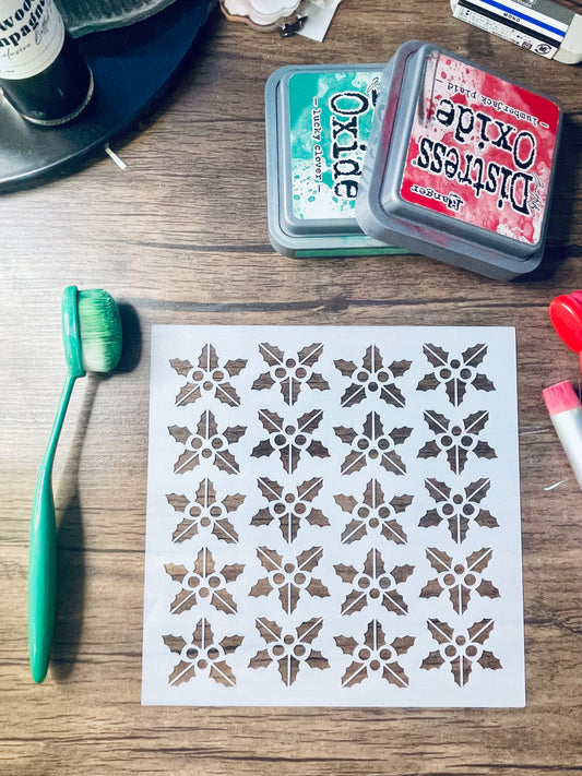 A sheet of stencils with a repeating snowflake or floral star motif, laid on a wooden surface next to a green-handled brush and two Distress Oxide ink pads.
