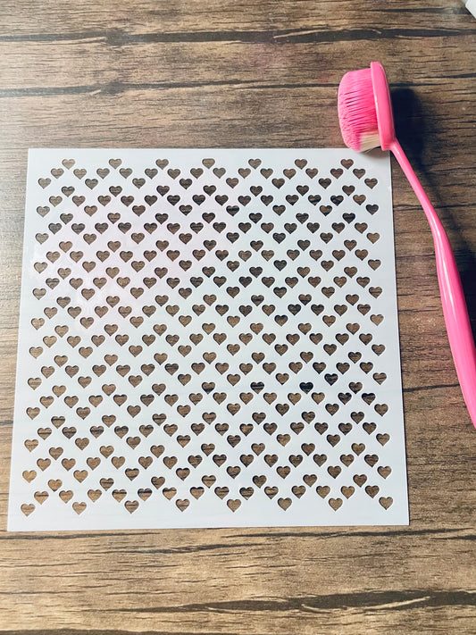 5.75” x 5.75” frosted stencil with Silhouettes of: tiny hearts horizontally lined up in a roughly zig zag shaped rows.  Stencil is on a wood surface with a pink brush 