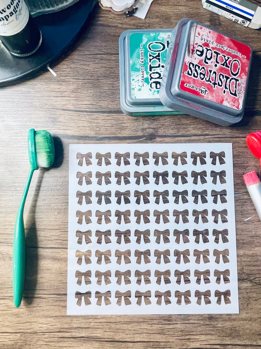 A sheet of small bow-shaped stickers or decals laid on a wooden surface, surrounded by craft supplies including two Distress Oxide ink pads, a green-handled brush, and other tools.