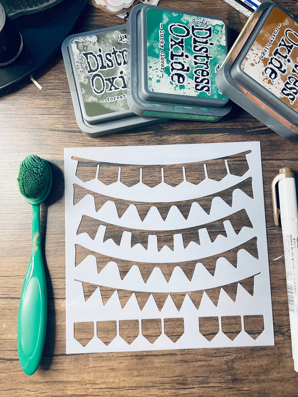 A white stencil sheet with multiple curved banner cutouts lies on a wooden surface, surrounded by craft supplies including a green-handled brush, Distress Oxi ink pads, and a white marker.