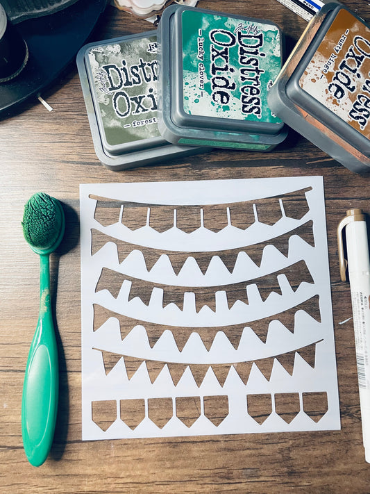 A white stencil sheet with multiple curved banner cutouts lies on a wooden surface, surrounded by craft supplies including a green-handled brush, Distress Oxi ink pads, and a white marker.