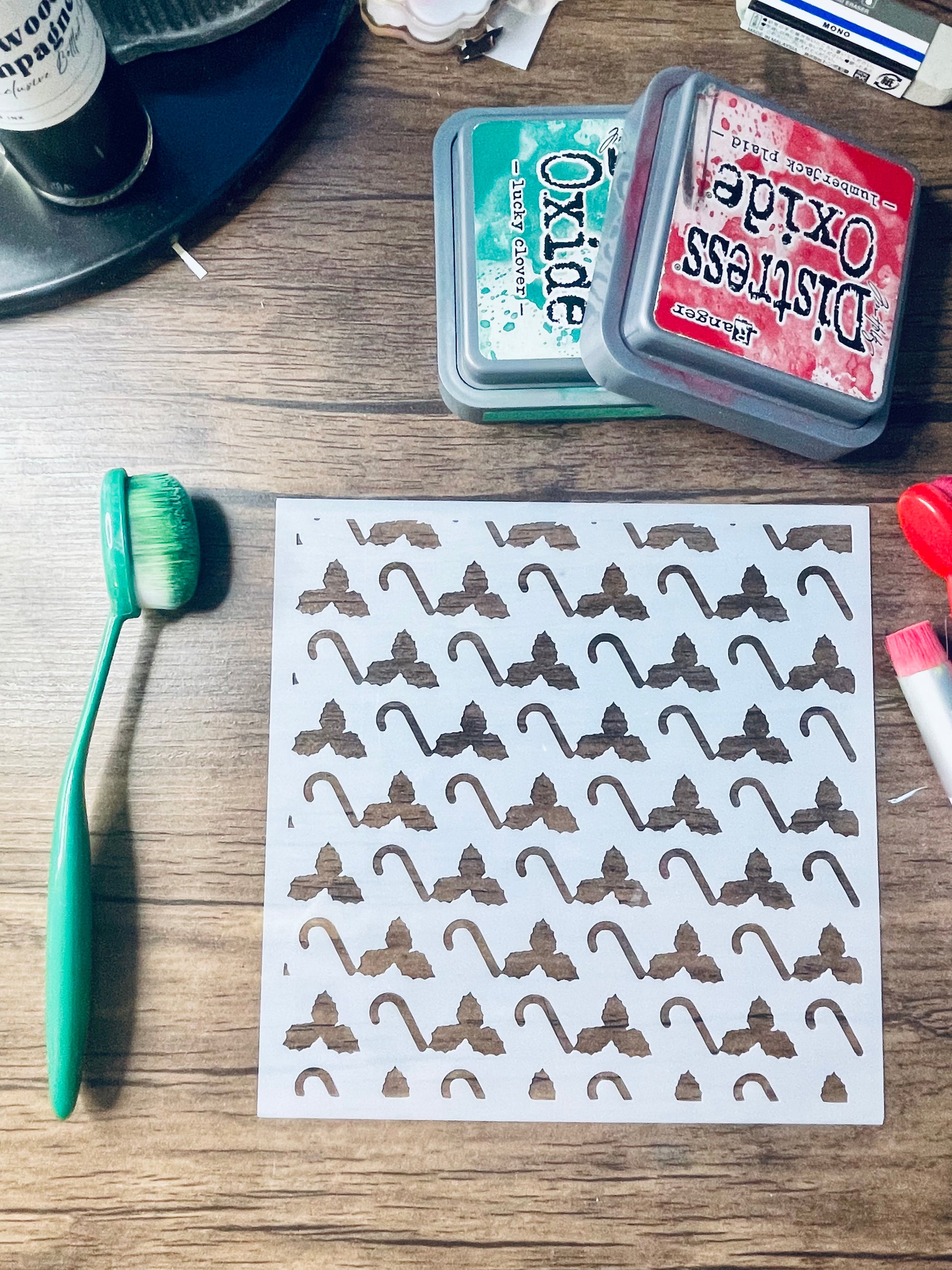 A sheet of white decals or stickers printed with a repeating brown bow-tie pattern, laid on a wooden tabletop alongside a green-handled brush, two Distress Oxide ink pads, and other small craft items.