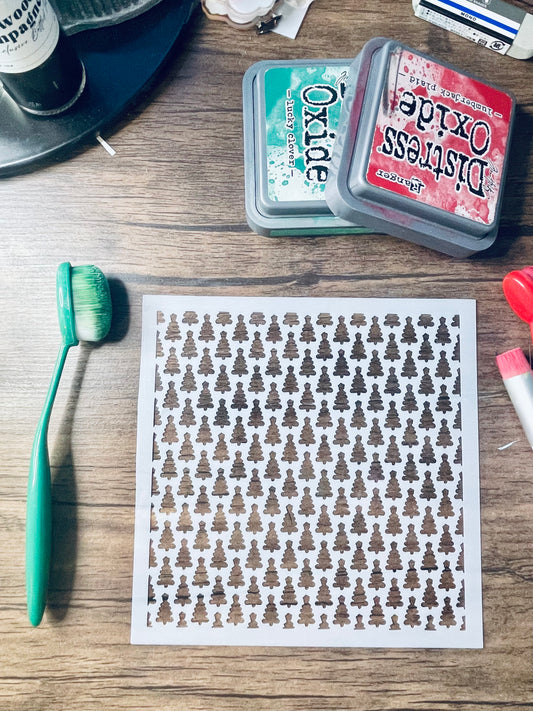 A square white sheet with a repeating Christmas tree stencil pattern, displayed on a wooden desk alongside craft supplies including an orange-handled brush, Distress Oxide ink pads, and other crafting tools.