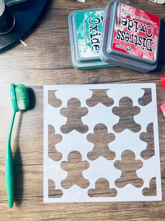 A white square stencil sheet with repeated gingerbread man cutouts, placed on a wooden surface alongside a green-handled brush and two Distress Oxide ink pads.