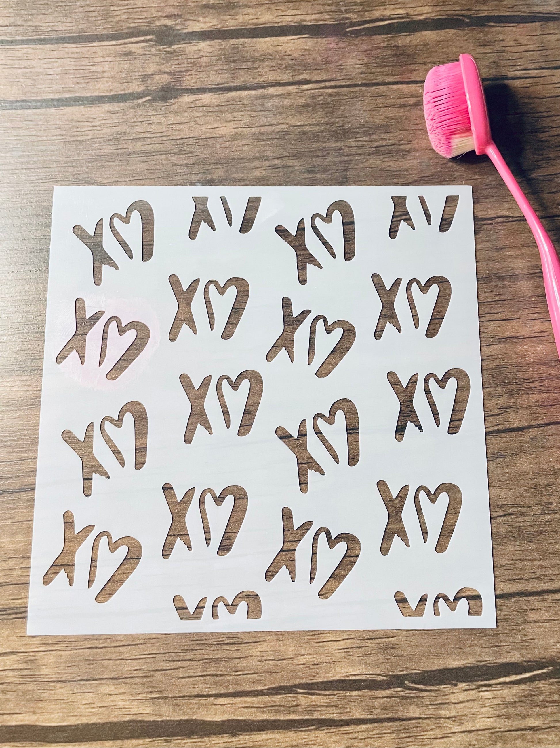 5.75” x 5.75” frosted stencil with Silhouettes of: XO in a grafiti type font in pattern around the stencil. The Os are hearts instead of Os.  Stencil is on a wood surface with a pink brush 