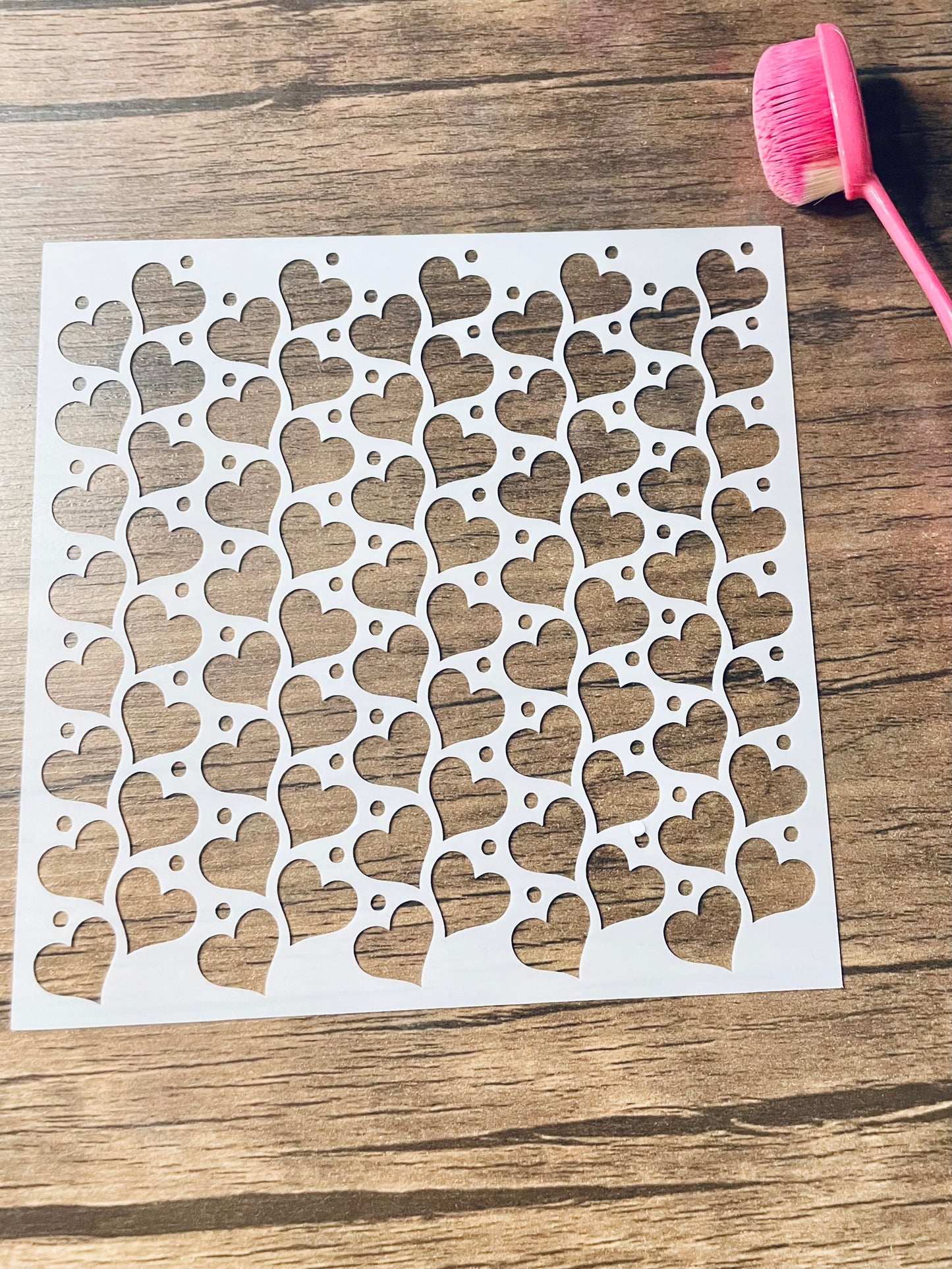 5.75” x 5.75” frosted stencil with Silhouettes of: Hearts tilted right and left in lines on a wooden surface with pink brush 