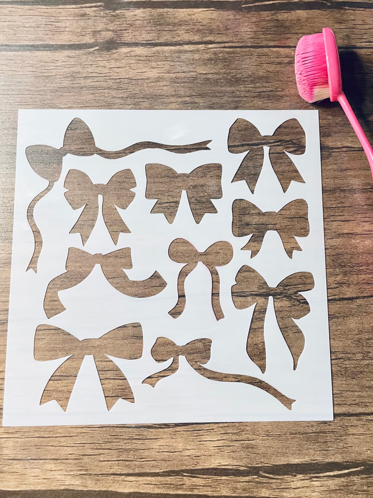 5.75” x 5.75” frosted stencil with Silhouettes of: ten different bows in various shapes and styles.  Stencil is on a wood surface with a pink brush 