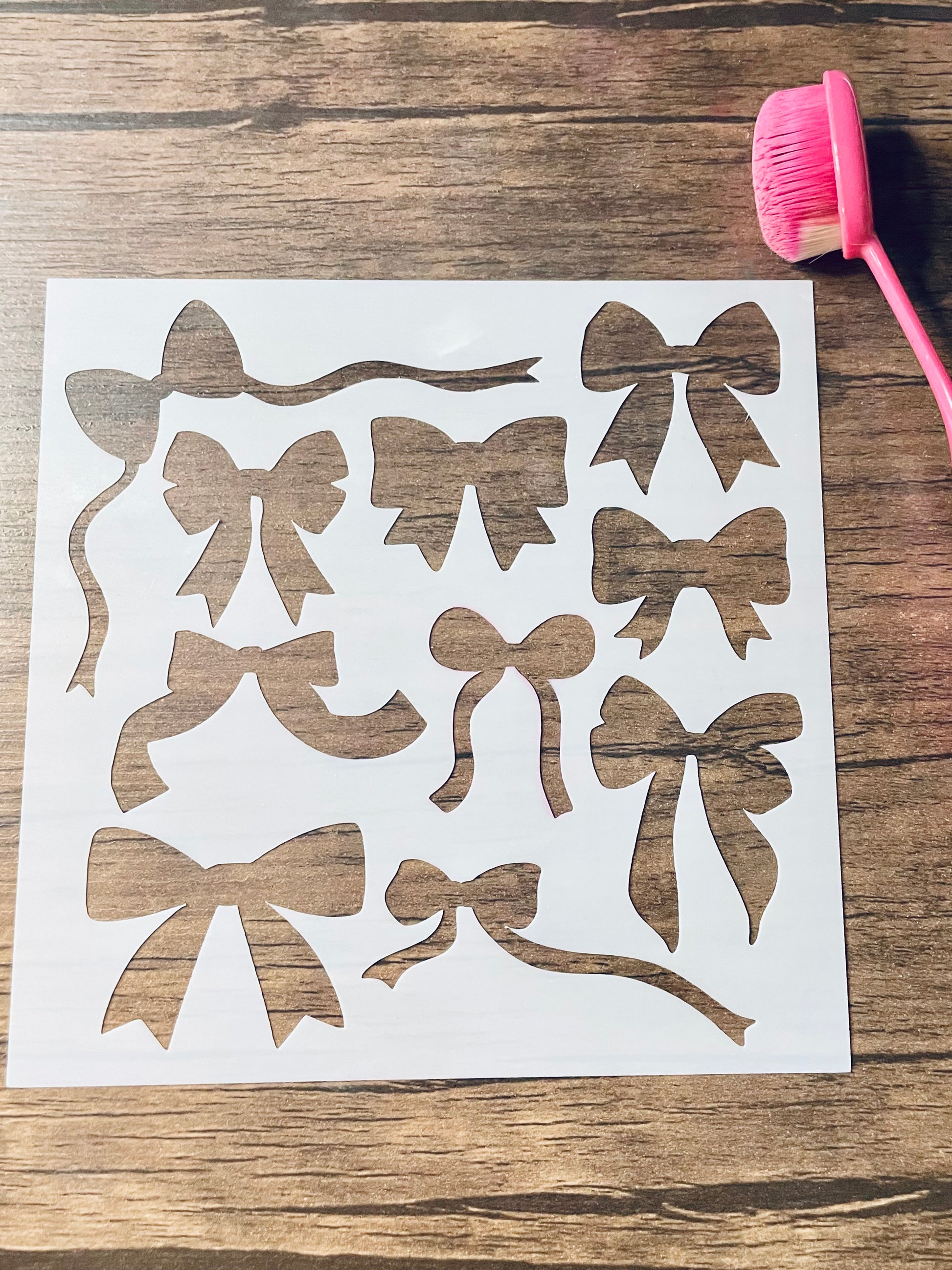 5.75” x 5.75” frosted stencil with Silhouettes of: ten different bows in various shapes and styles.  Stencil is on a wood surface with a pink brush 