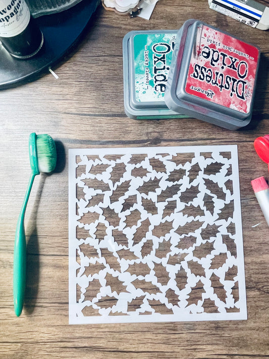 A square white stencil sheet with an intricate repeating geometric cutout pattern, displayed on a wooden tabletop alongside craft supplies including an orange-handled brush, Distress Oxide ink pads, and other crafting tools.