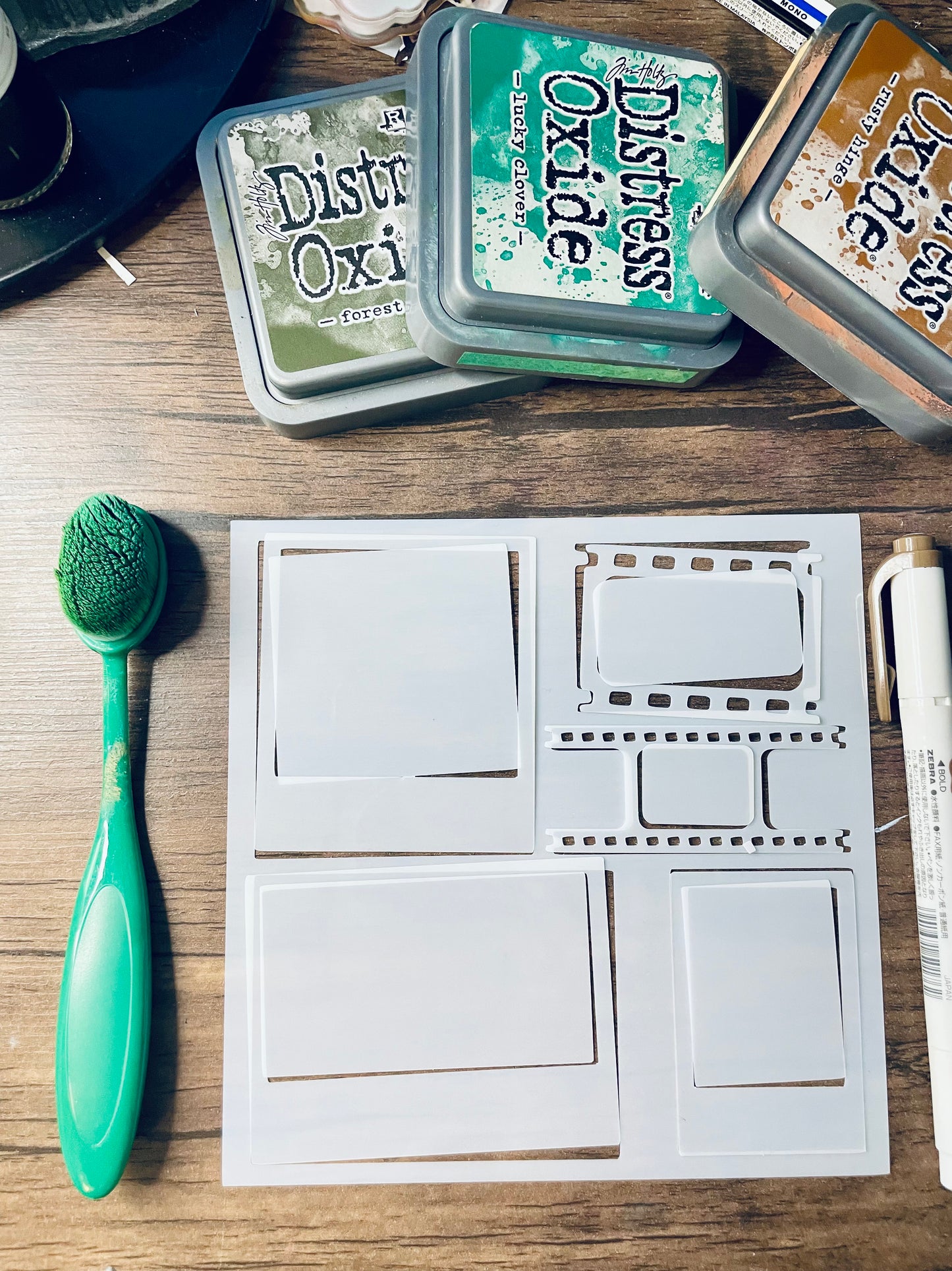 A white metal die-cut stamping plate with multiple template shapes is displayed on a wooden surface, accompanied by a green-handled brush, a white marker, and three Distress Oxi ink pads in green, teal, and brown.